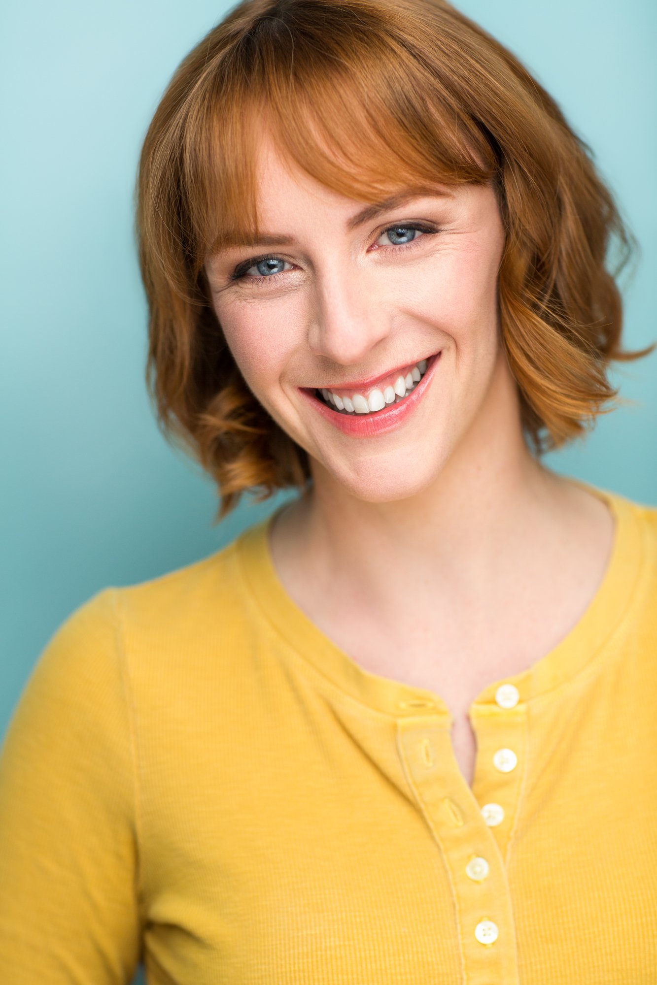 Smiling headshot of a red-haired woman wearing a yellow top against a bright blue background.