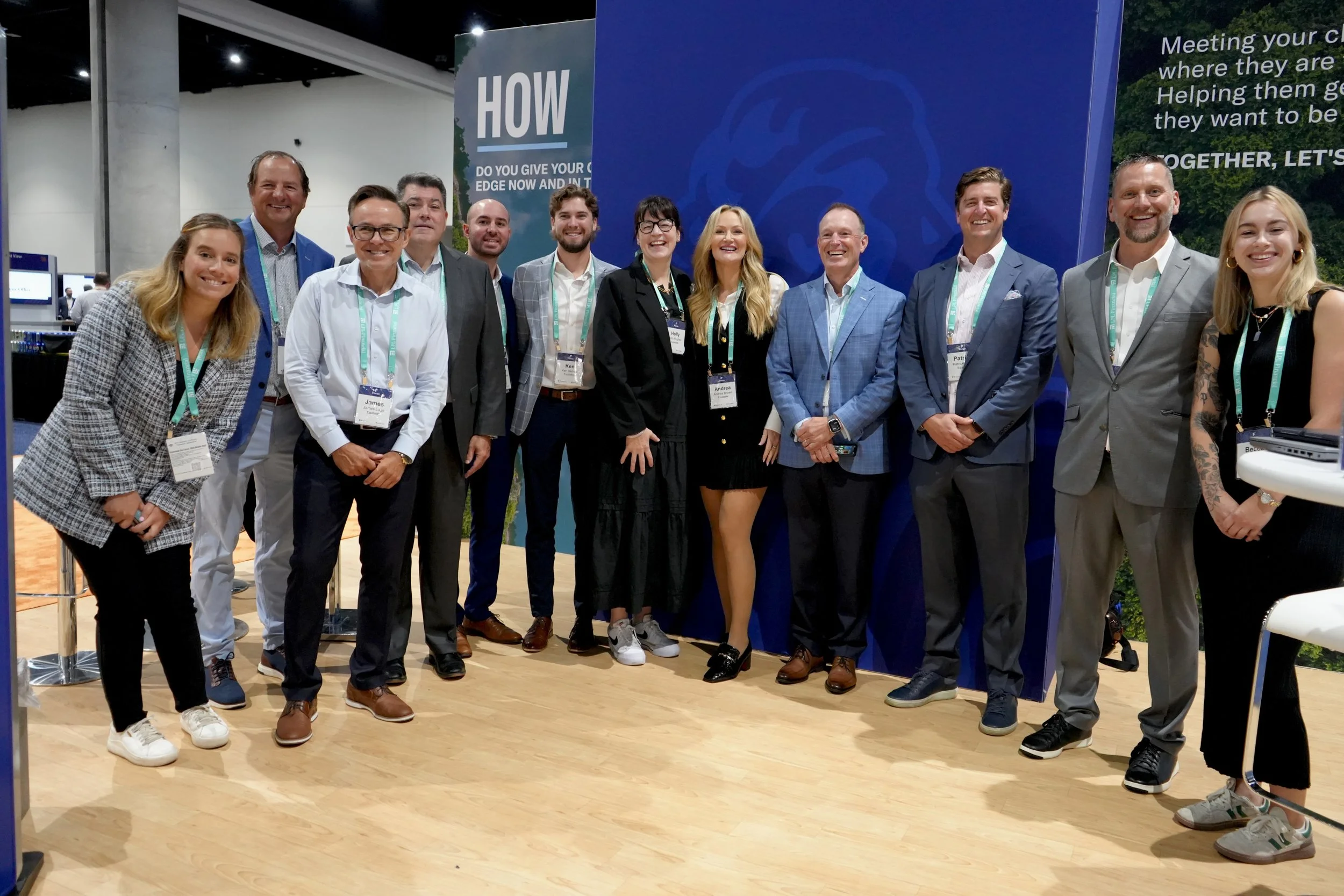 A group of 12 people dressed in business casual attire standing together at a conference or event, smiling for the camera in front of a blue backdrop.