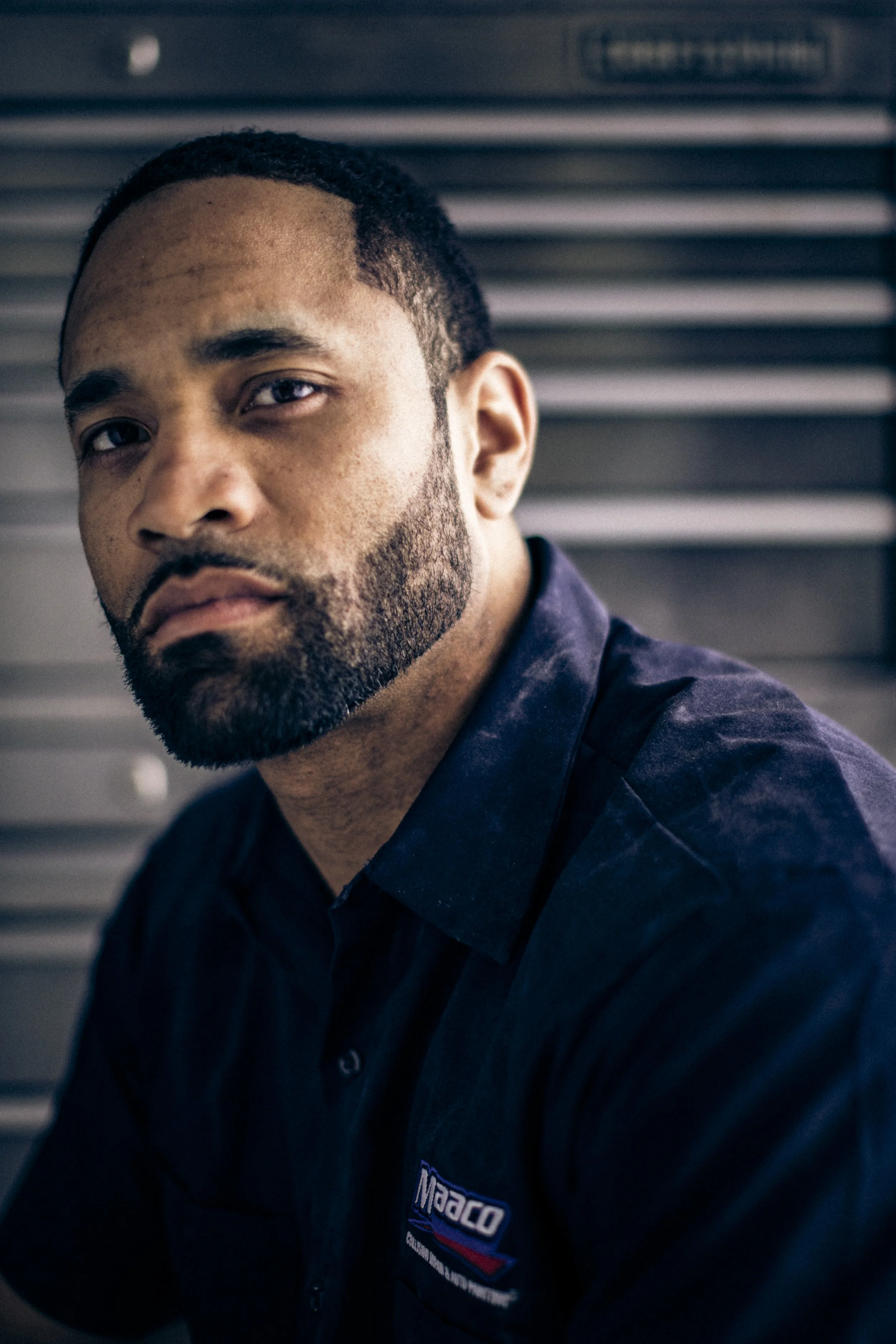 Close-up of a man with a beard and short hair, wearing a black work shirt with a logo, looking serious, with a metal background.