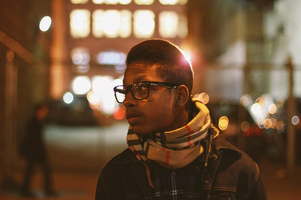 A young man with glasses and a scarf standing on a city sidewalk at night, with blurred city lights in the background.