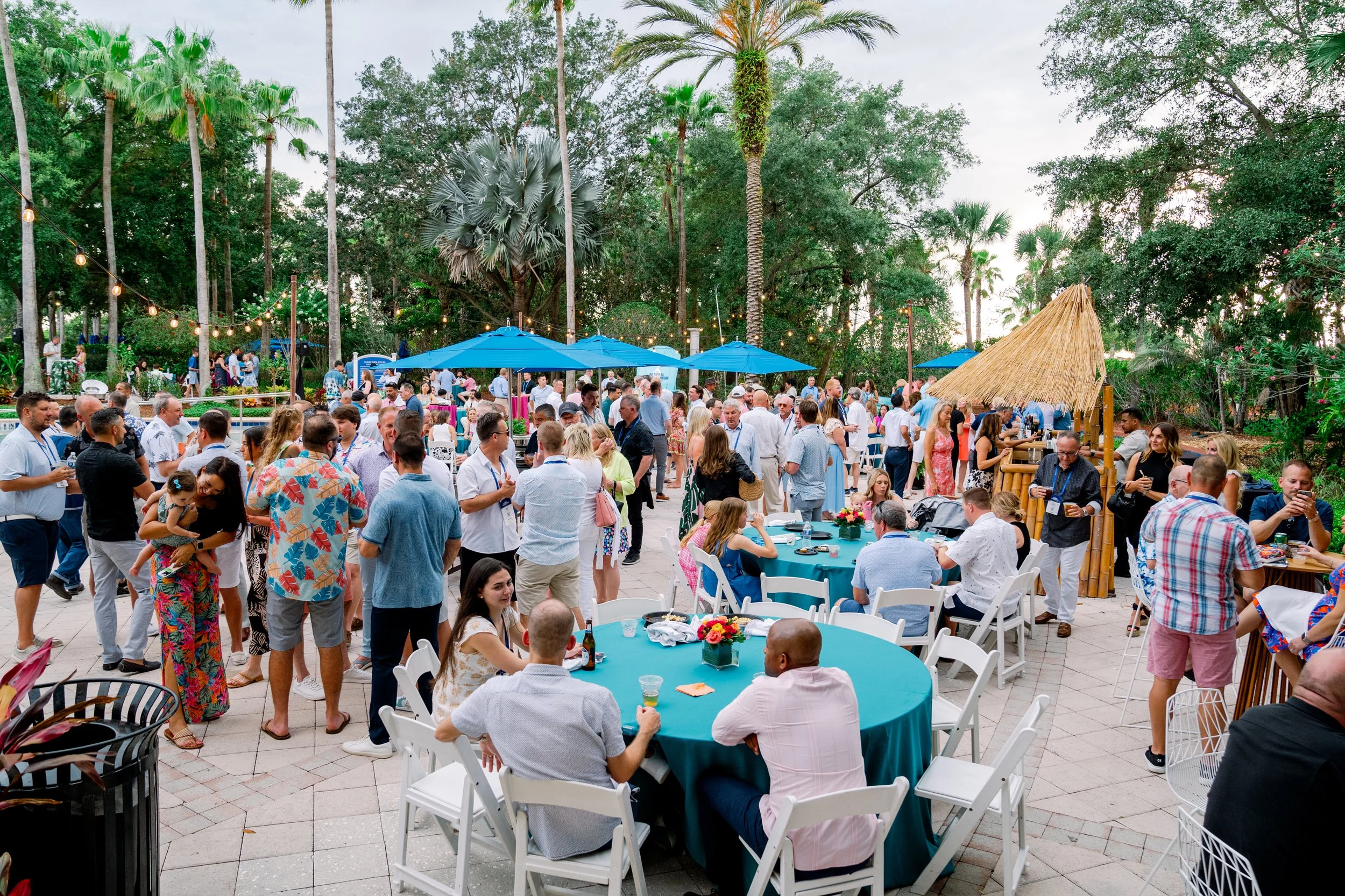 Outdoor social gathering or party with many people around tables and under umbrellas, surrounded by trees and string lights.