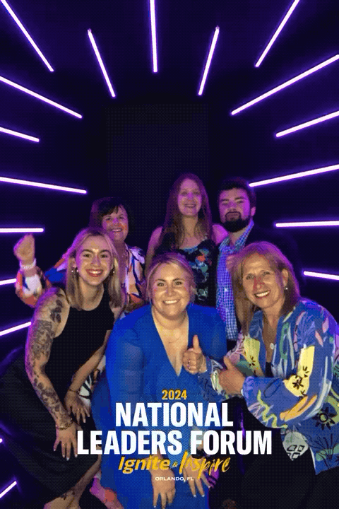 Group of six people smiling and posing in front of a neon light display at the 2024 National Leaders Forum in Orlando, Florida.
