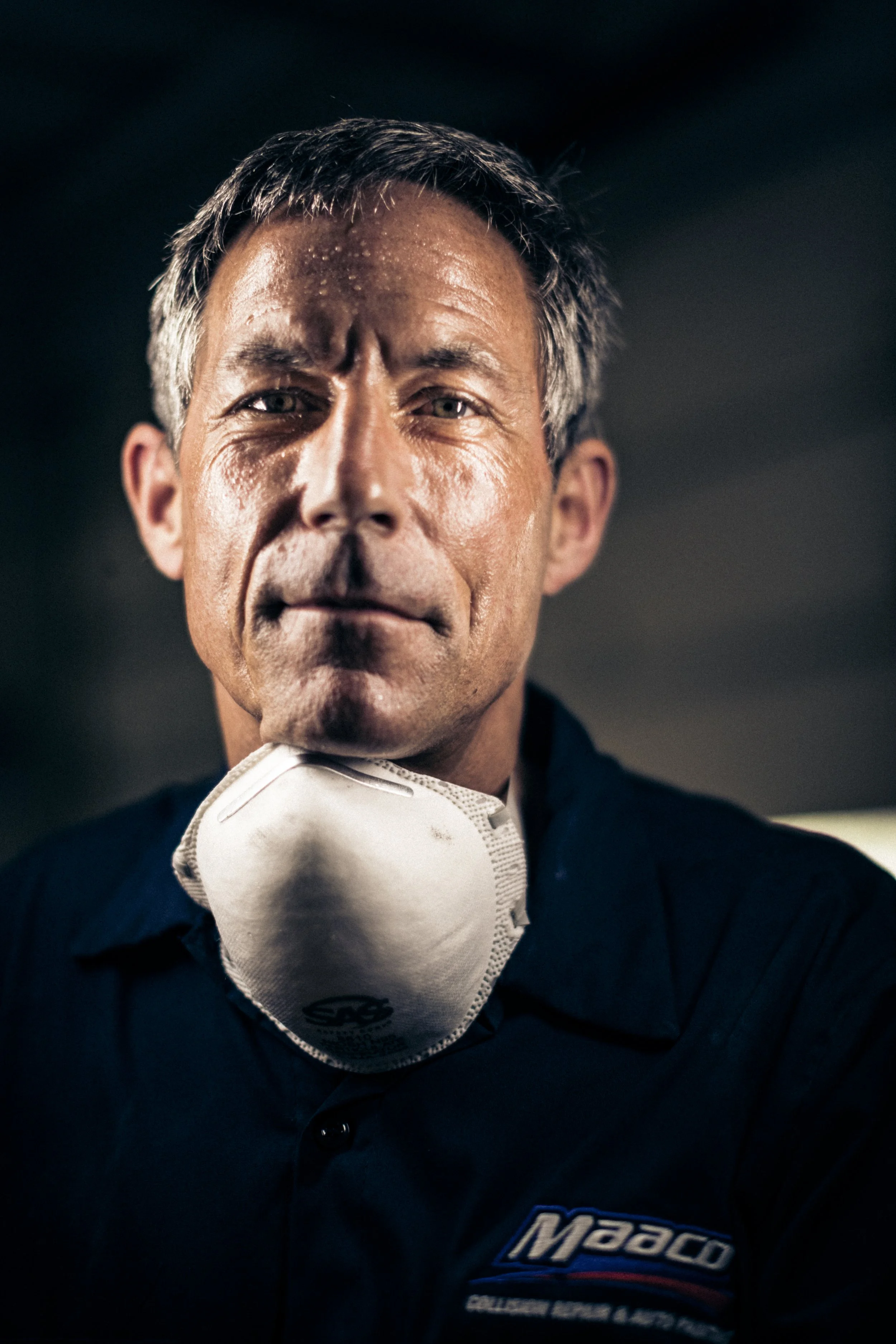 A man with gray hair and a serious expression wearing a dark shirt with a logo that says 'Maaco' and a white mask hanging around his neck.