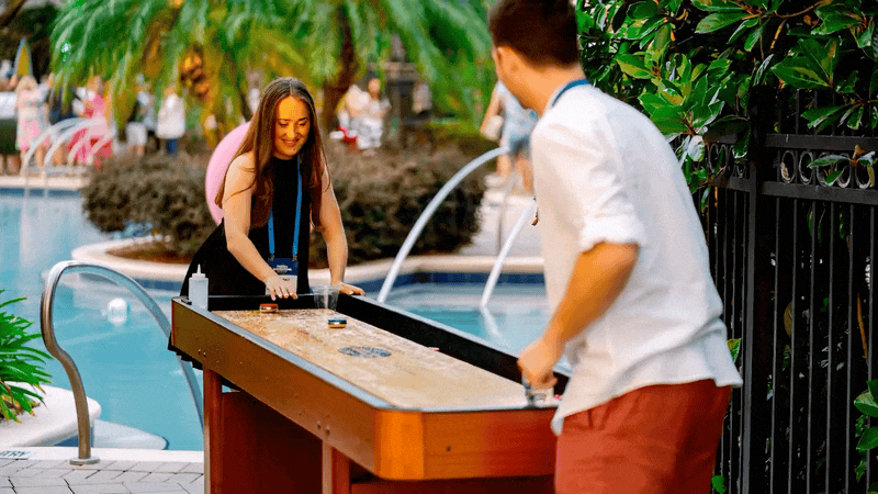 Two people playing shuffleboard by a swimming pool, surrounded by tropical plants and lounge chairs.