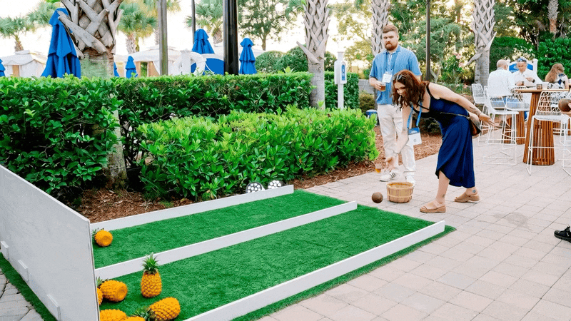 A woman plays mini golf on an outdoor course, with a man watching nearby. The course has a small green turf area with pineapple and flower decorations, and is bordered by bushes and trees. There are other people sitting at tables in the background.