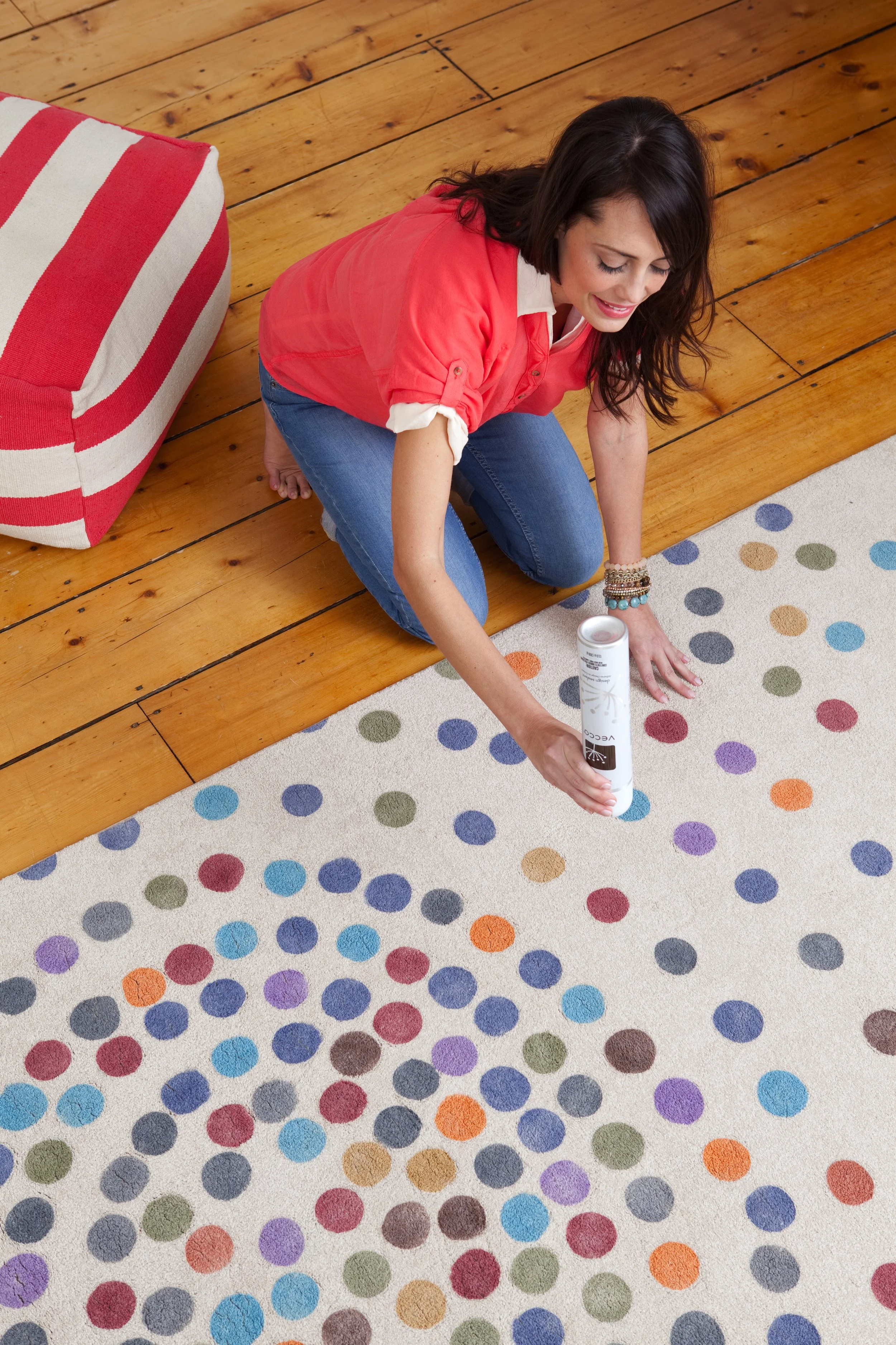 A woman kneeling on a wooden floor, holding a can of carpet cleaner, working on a colorful polka dot rug.