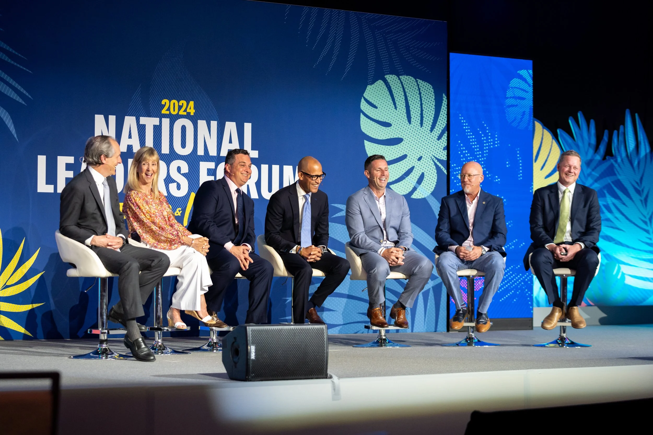 A panel of seven people sitting on a stage at the 2024 National Leaders Forum, smiling and engaged in a discussion, with a blue backdrop featuring the event name and decorative leaf patterns.