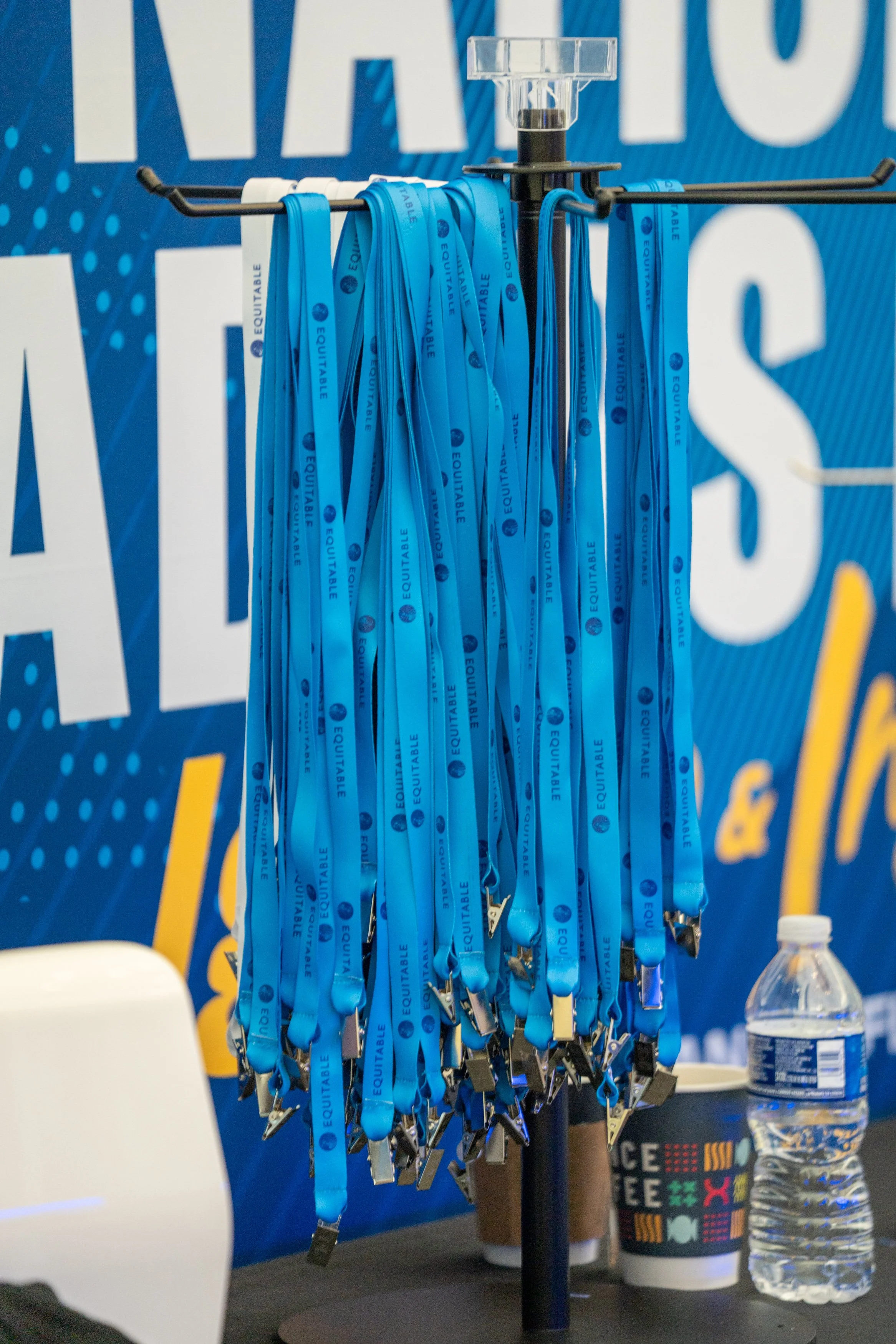 Blue event lanyards hanging on a black stand, with a water bottle and paper cup on a table in the background.