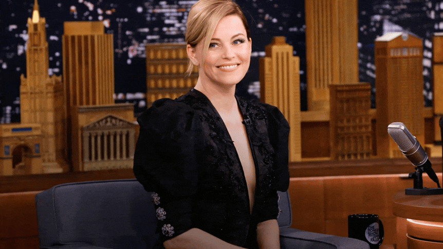 A woman with blonde hair in a black dress sitting on a talk show set with a city skyline backdrop and a microphone on the table.