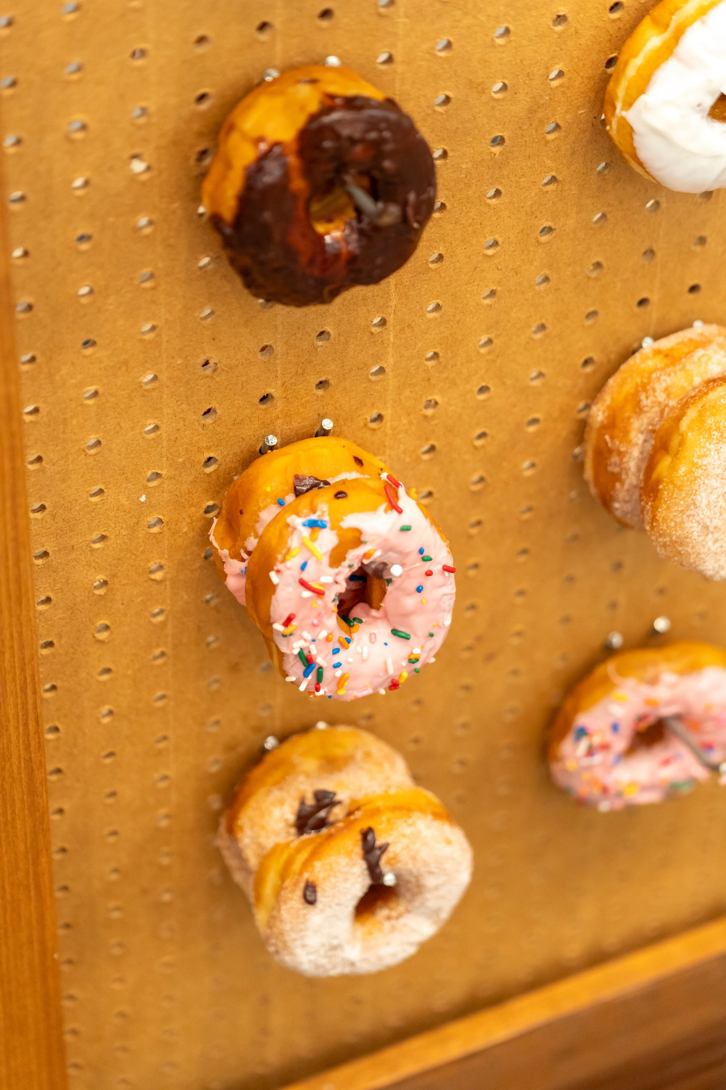 Assorted mini donuts with different toppings including chocolate, pink frosting with sprinkles, and powdered sugar, displayed on a wooden board.