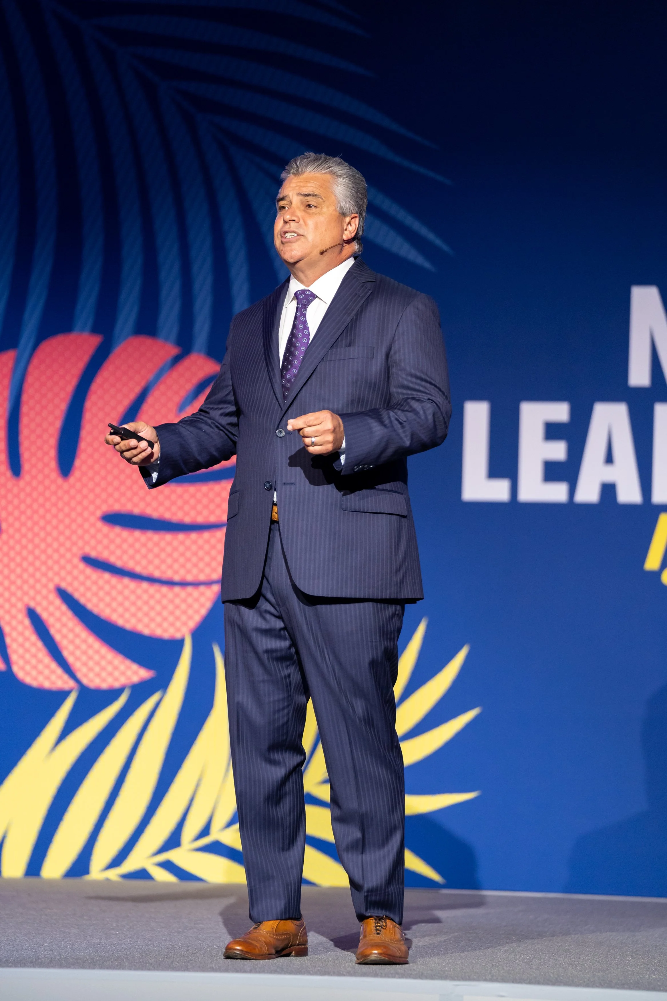 A man in a navy pinstripe suit giving a presentation on stage, holding a remote, with a large colorful backdrop behind him.