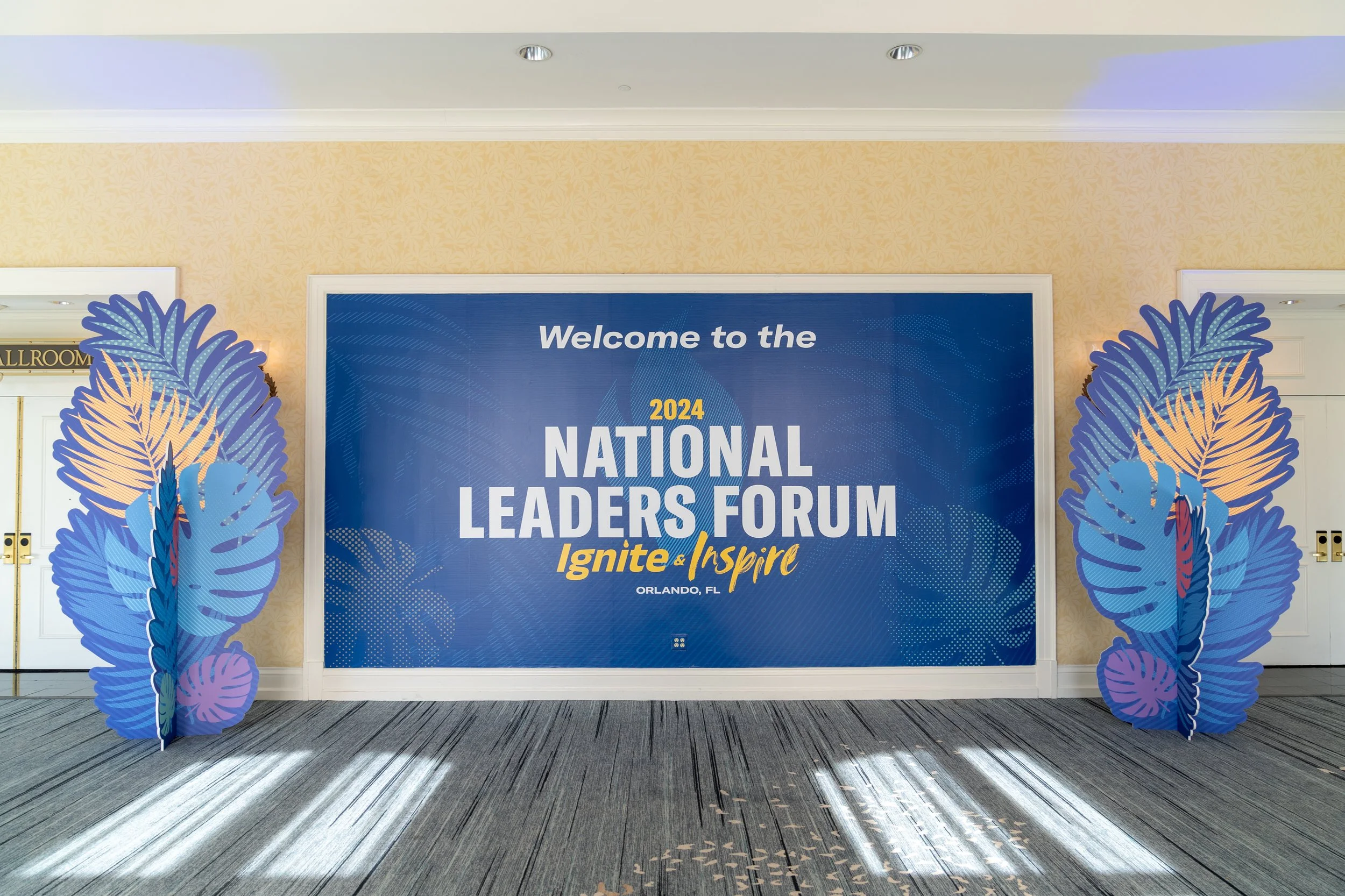 A large blue banner with white and yellow text reads "Welcome to the 2024 National Leaders Forum Ignite & Inspire Orlando, FL". There are decorative cutouts of large tropical leaves on each side of the banner, with a patterned carpet floor and cream-
