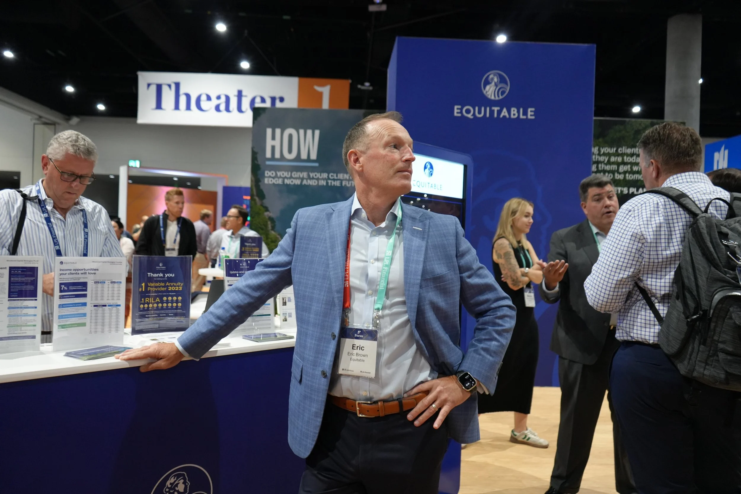 A man in a light blue blazer and white shirt standing with his hand on his hip and the other hand resting on a booth table at a conference or expo, with other people talking and browsing around, and large blue and orange signs in the background.