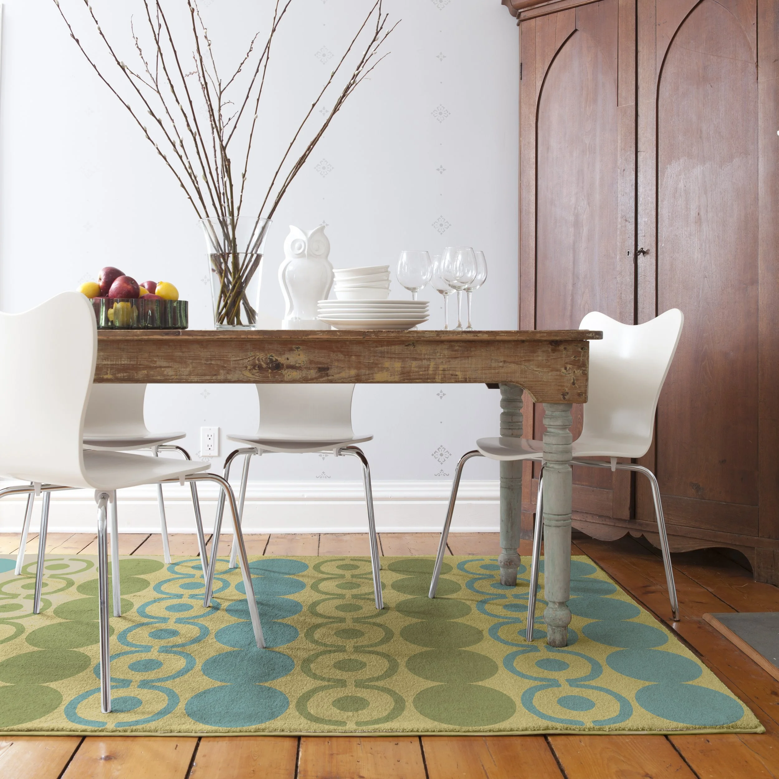 A dining room with a wooden table, white modern chairs, a green and blue patterned rug, and a wooden cabinet in the background. The table has a vase with branches, a bowl of fruit, white plates, a ceramic owl, and three wine glasses.