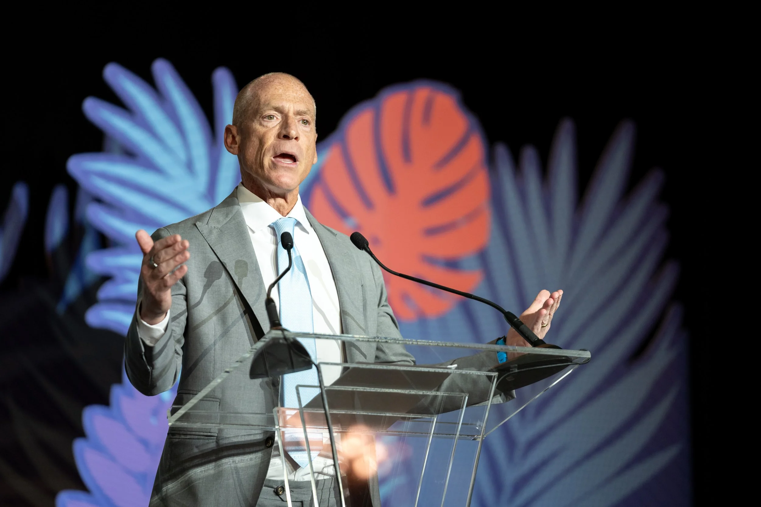 A man in a gray suit and light blue tie speaking at a podium with microphones, gesturing with his hands, against a dark background with stylized blue and orange leaf-like shapes.