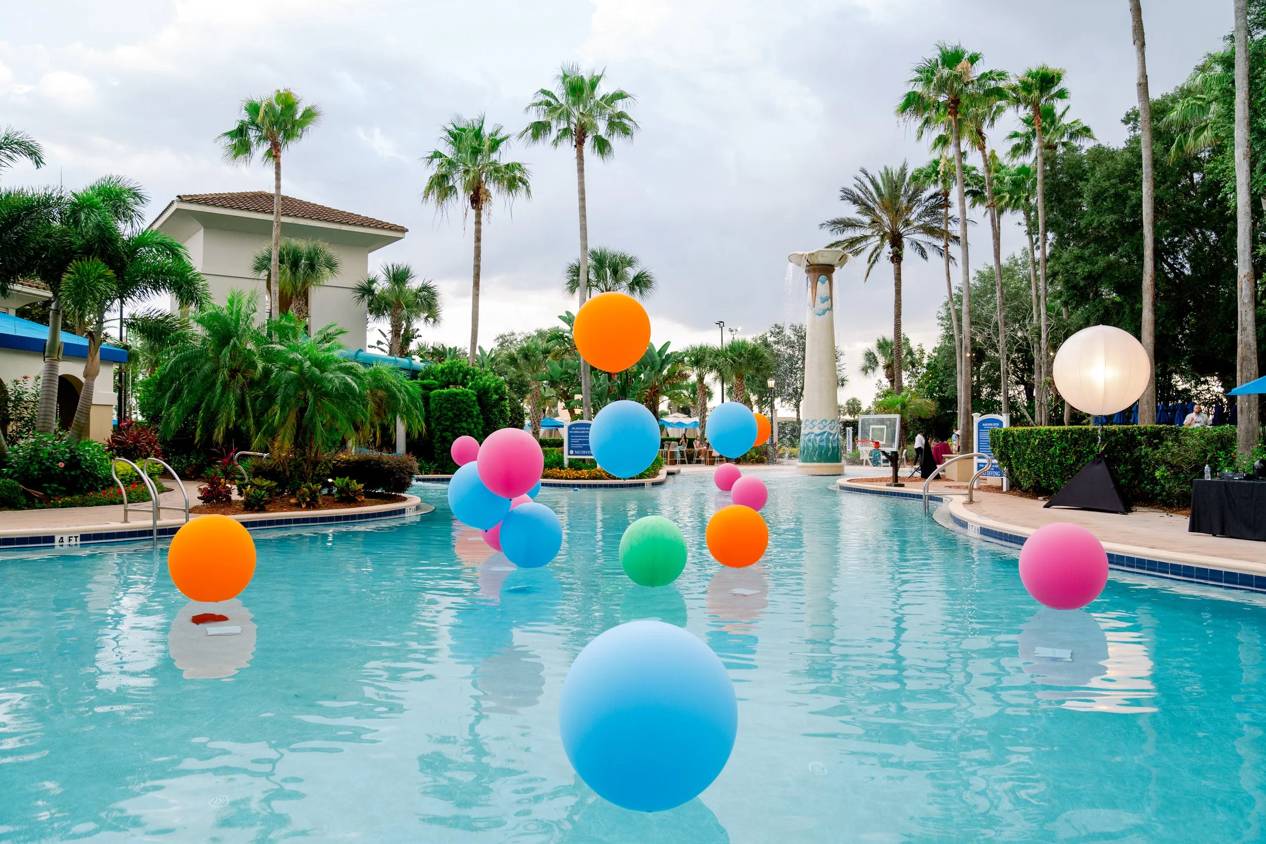 Colorful balloons floating in a swimming pool at a tropical resort with palm trees, a fountain, and lounge areas.