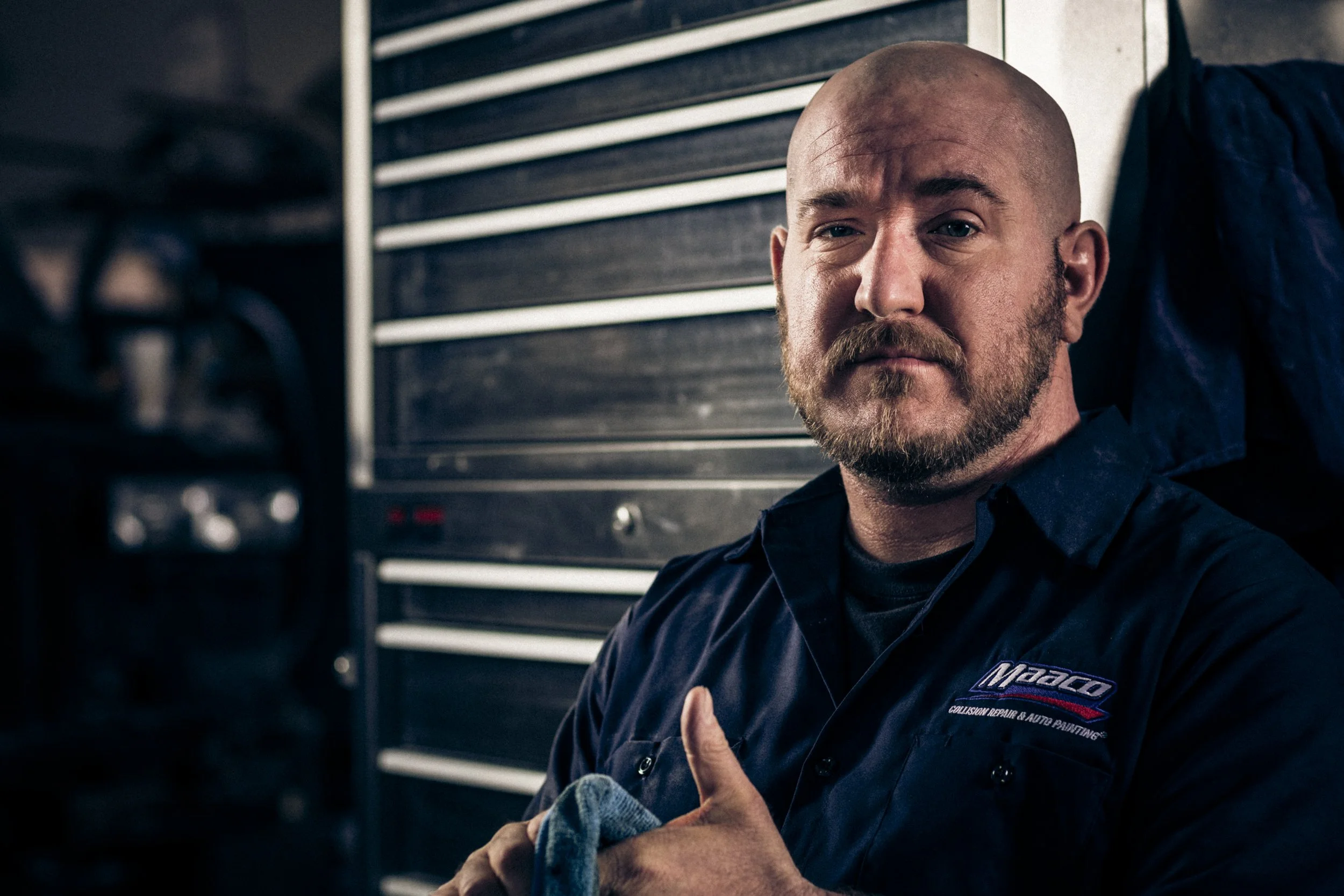 A man with a bald head and beard, wearing a dark work shirt, is sitting in a garage or workshop, giving a thumbs-up. There are tool drawers in the background.