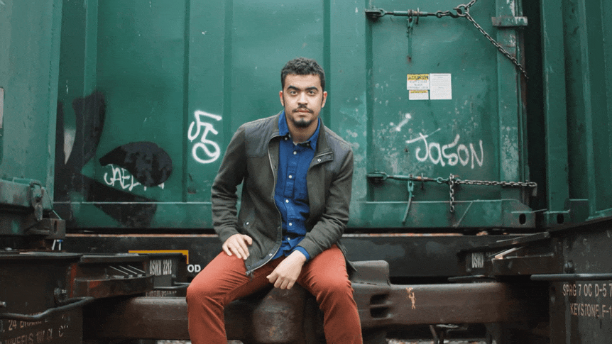 A young man with dark hair, a dark beard, wearing a dark jacket over a blue shirt and reddish-brown pants, sitting on a train car with graffiti in an industrial setting.