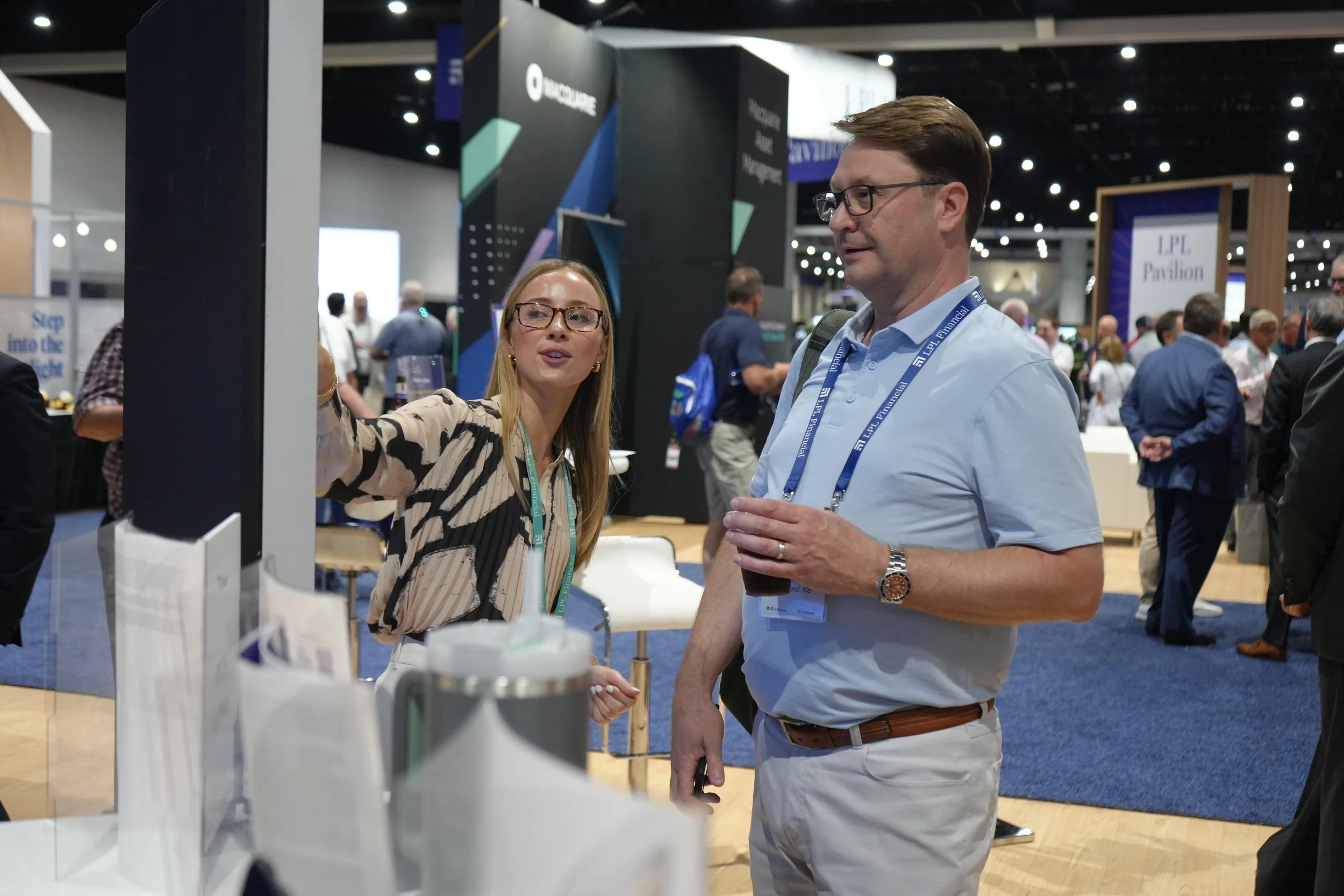 A woman is explaining something at a booth to a man with glasses in a crowded convention center.