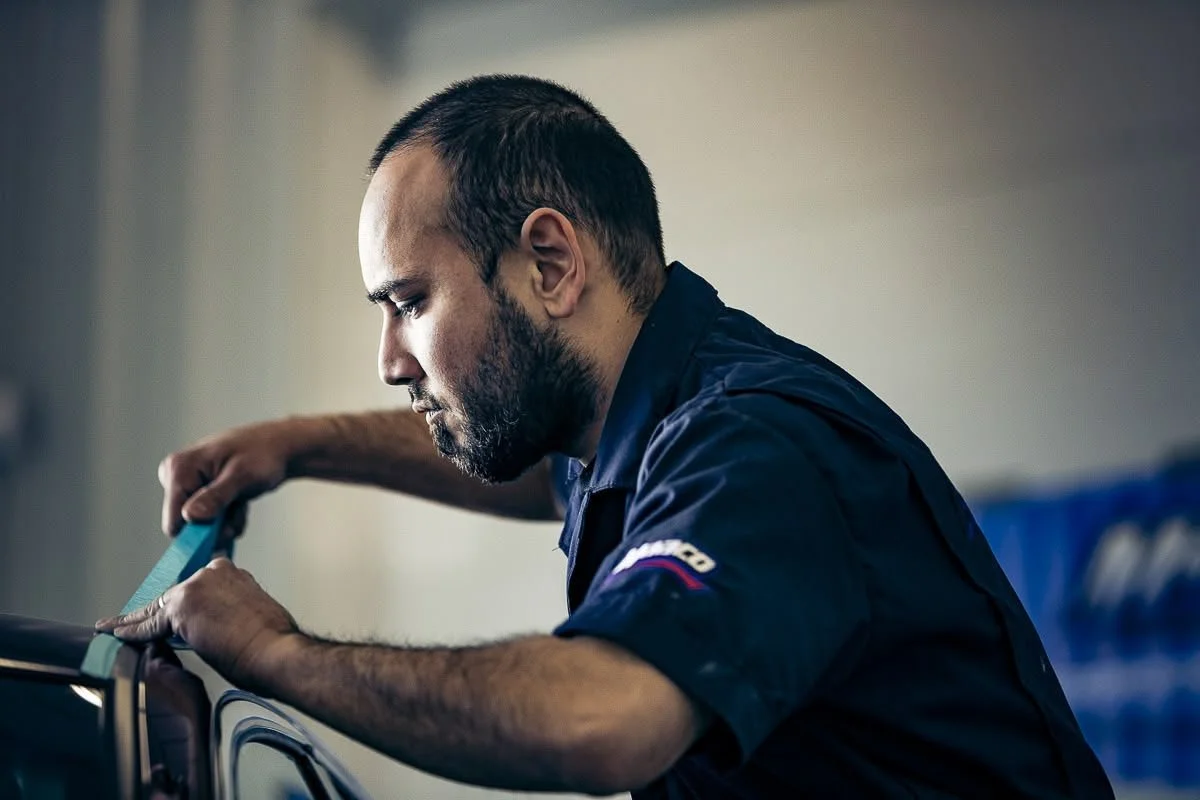 A man in a blue shirt is working on a vehicle, applying a blue strap or tape to its surface, with a focused expression.