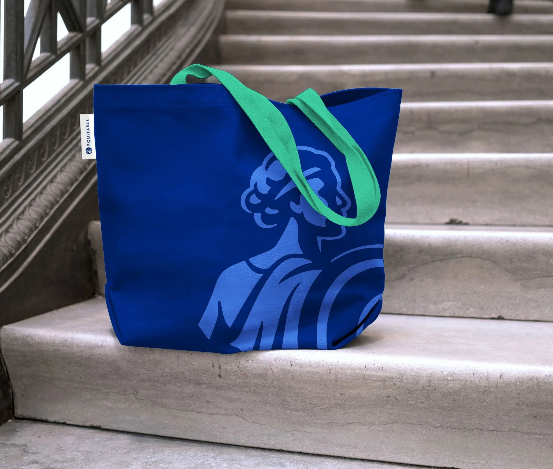 A blue tote bag with green handles sitting on a stone staircase, featuring a light blue illustration of the Statue of Liberty's head and torch.