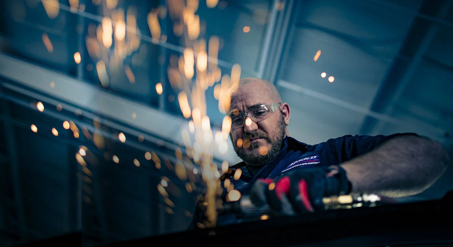 A man wearing safety glasses, a work uniform, and gloves is welding or using a grinder, creating sparks in an industrial or workshop setting.