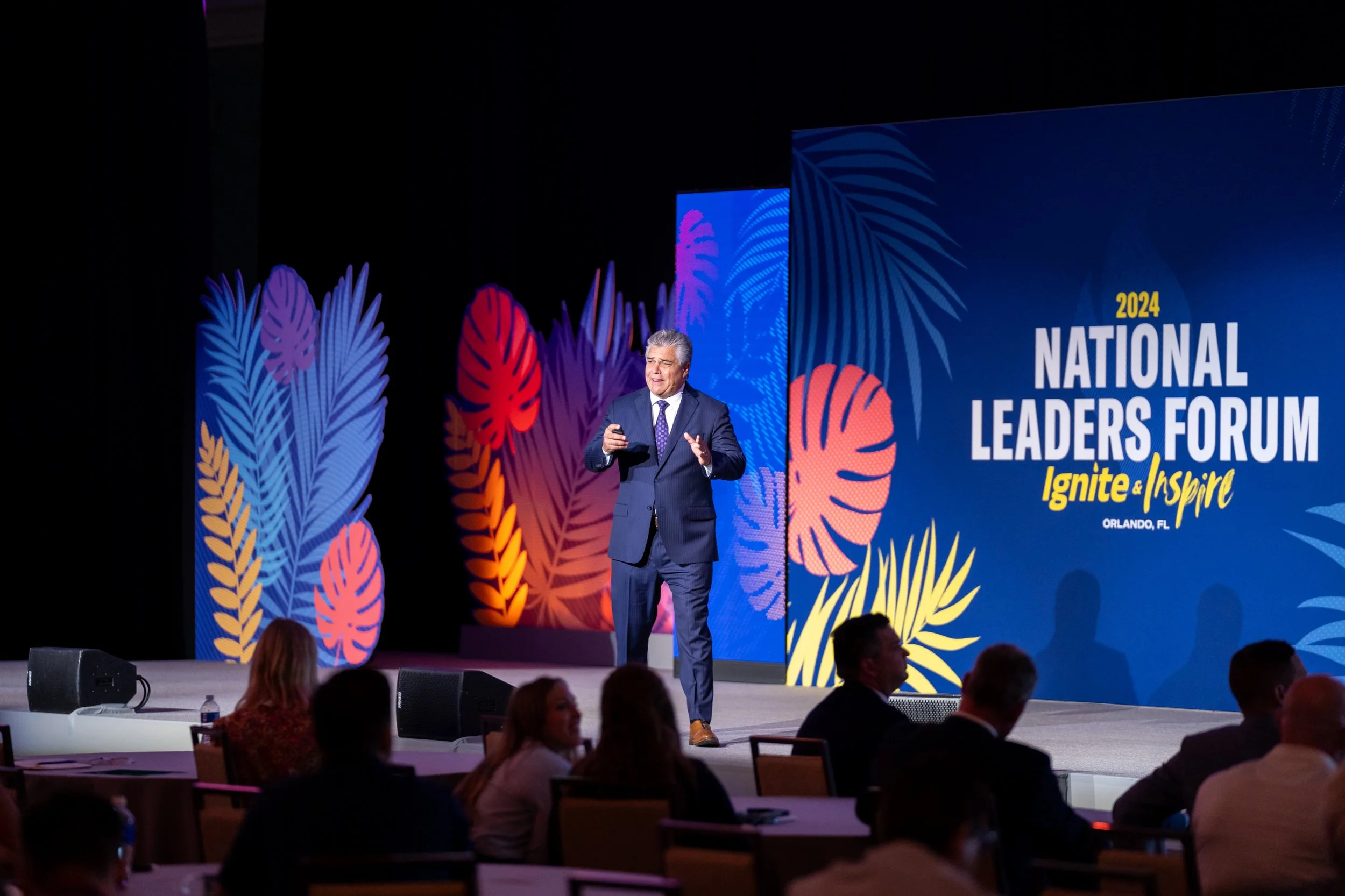A man in a suit giving a presentation at the 2024 National Leaders Forum in Orlando, Florida, with a colorful backdrop of leaves and the event's branding that includes the words 'Ignite & Inspire'.
