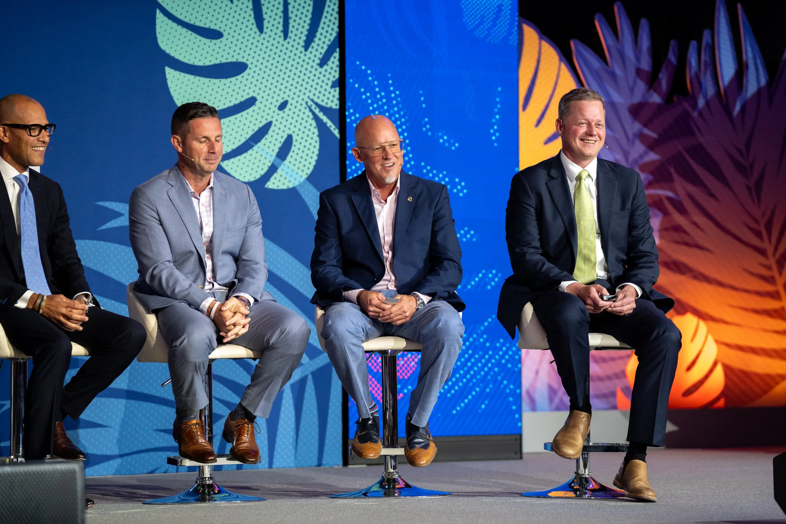 Four men in business attire sitting on stools on stage, smiling and engaging in a panel discussion, with vibrant blue and orange decorative backgrounds.