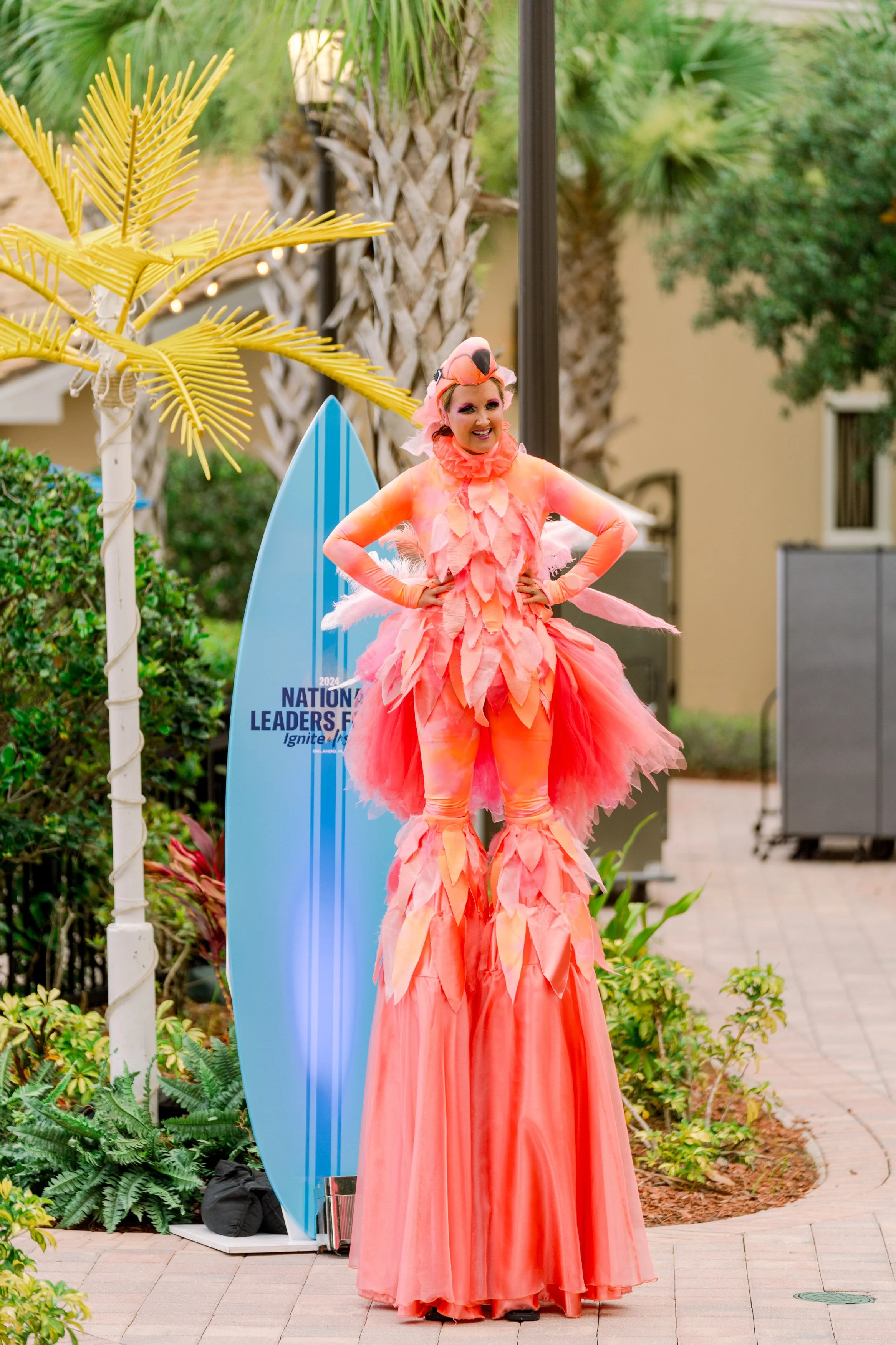 A performer dressed in a bright pink flamingo costume, standing on stilts, outdoors near a blue surfboard sign that reads 'National Leaders Ignite 2024'.