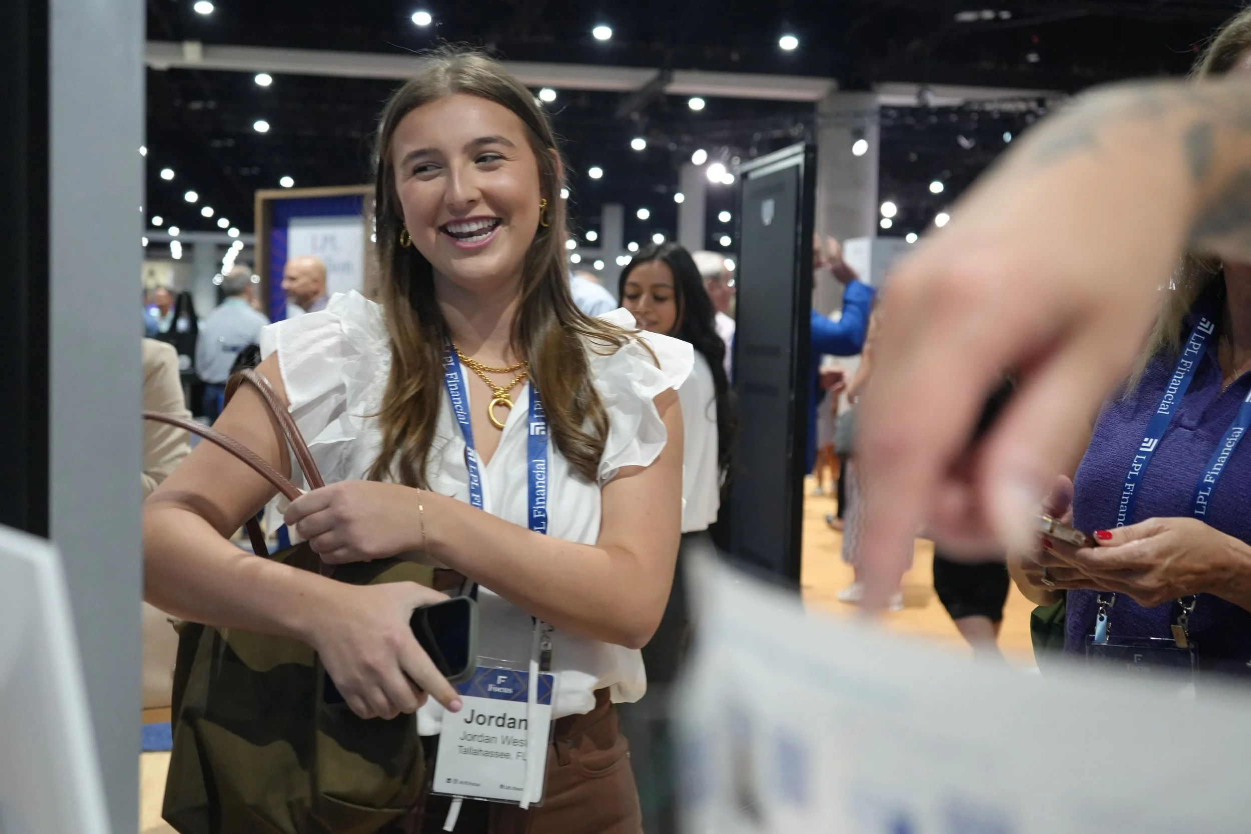 A young woman with long brown hair is smiling at a professional conference. She is wearing a white blouse with ruffled sleeves, gold jewelry, and a name badge. The background shows other attendees, conference booths, and bright lighting.