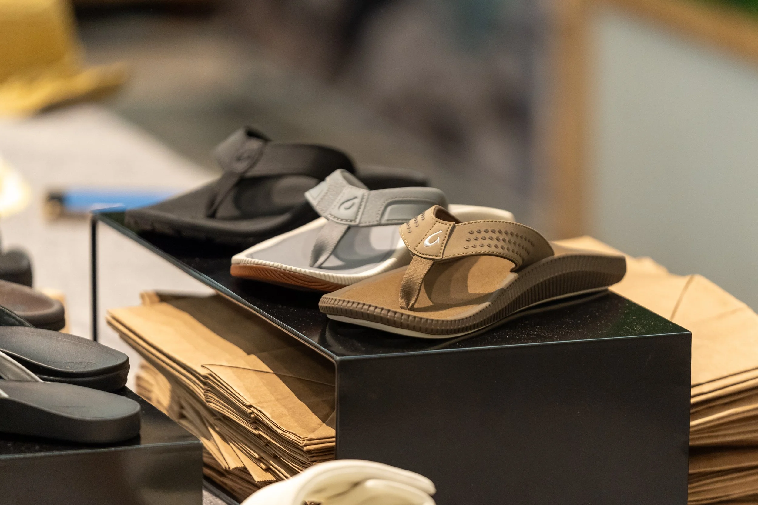 Display of three pairs of sandals in black, gray, and brown on a black platform in a retail store with paper bags underneath.