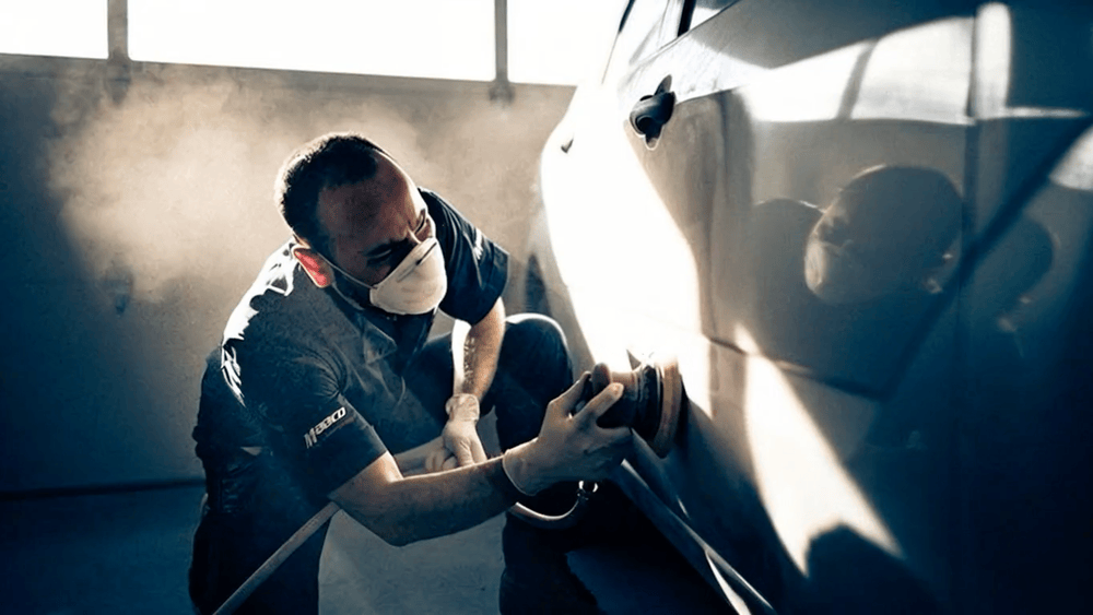 A person polishing or buffing a car's exterior with a power buffer, wearing a face mask, in a garage or workshop.