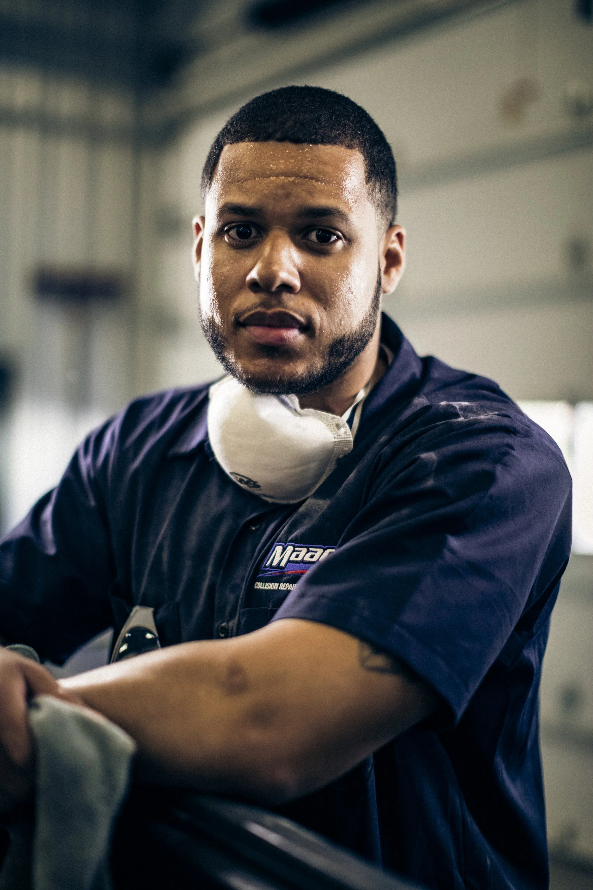 A young man with short dark hair and a beard, wearing a navy blue uniform with a logo that reads 'Moag Collision Repair', and a white mask around his neck, working in an auto repair shop.