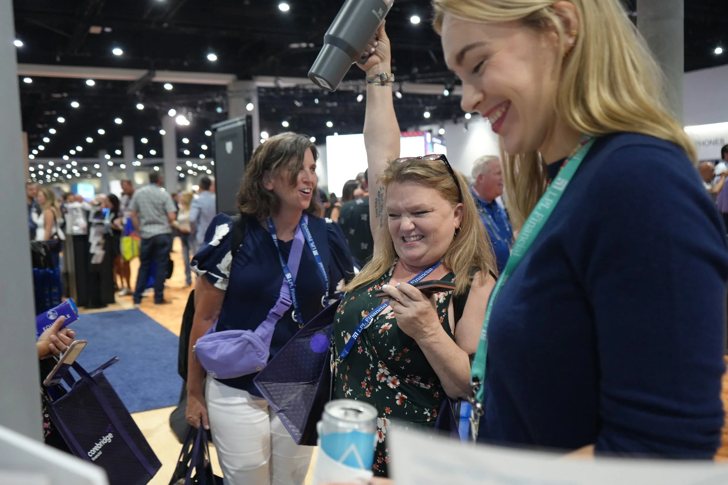 Three women at a trade show or conference, smiling and engaging with a smartphone, with other attendees in the background.