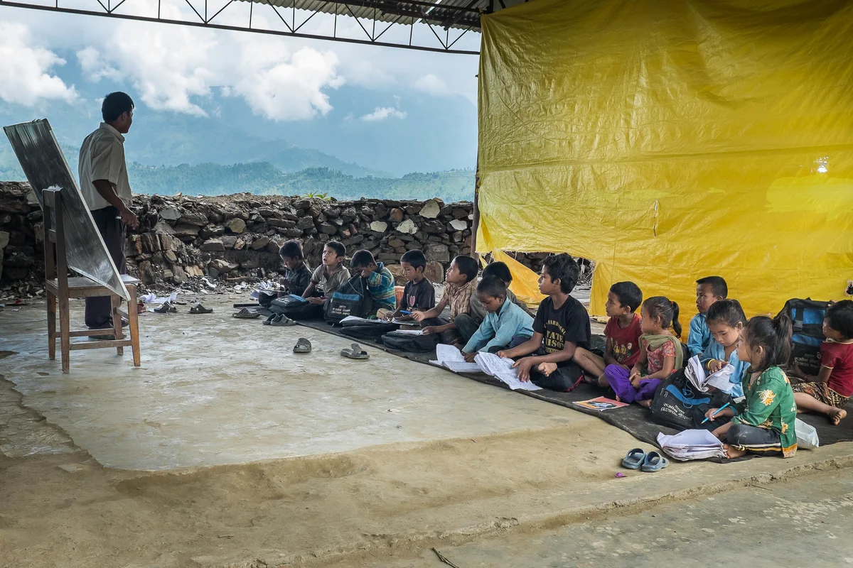 Children attend a class where their school once stood on 10 August 2015 in Sipa Pokhari. The school now only has one temporary learning center set up by NGOs, which has just two classrooms. The rest of the students attend classes underneath a metal rooftop, with no walls and only tarps to separate each classroom.