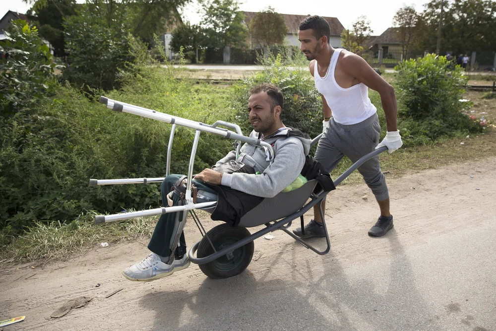Massur Nasser pushes his friend, Gazi El Fadour, in a wheelbarrow with a flat tire in Horgos, Serbia. El Fadour lost both his legs when ISIS attacked his university in Aleppo. The two men have been traveling companions since meeting months earlier in Turkey (Jodi Hilton/IRIN)