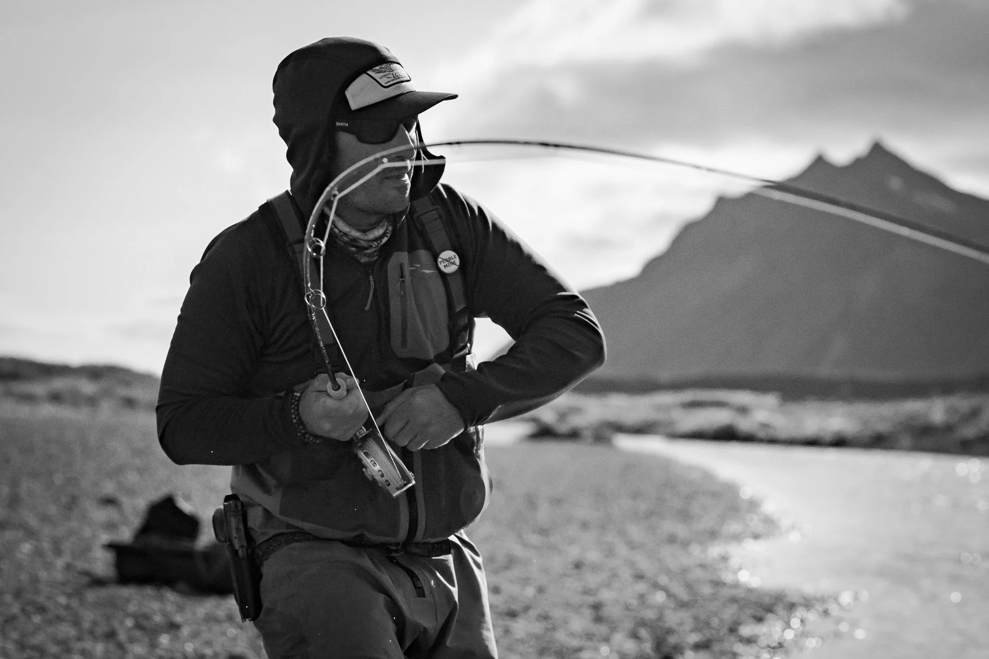 Fly fisherman casting beside an Alaskan river with mountains behind him