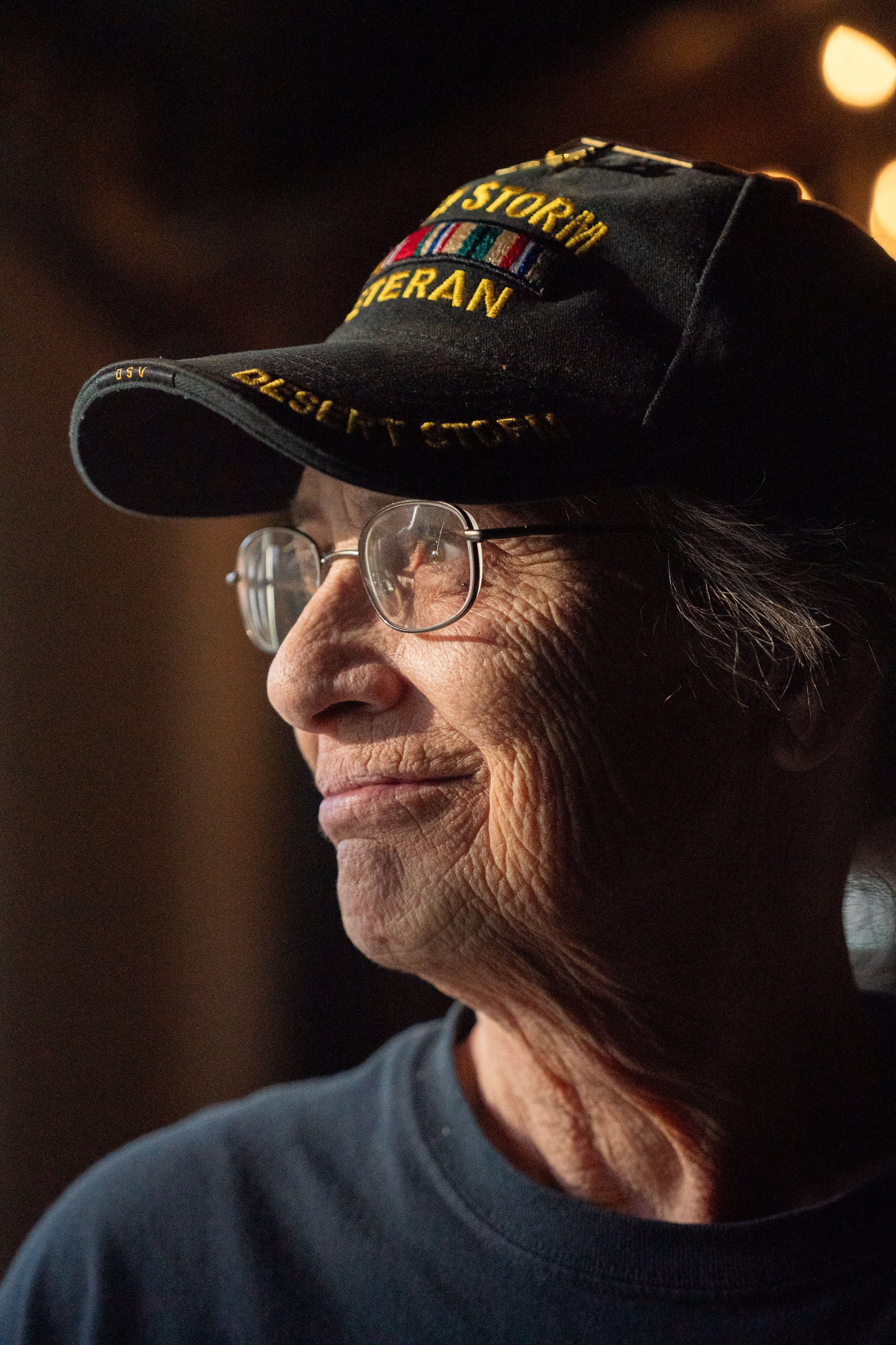 Side portrait of a veteran wearing glasses and a Desert Storm Veteran cap in warm indoor light