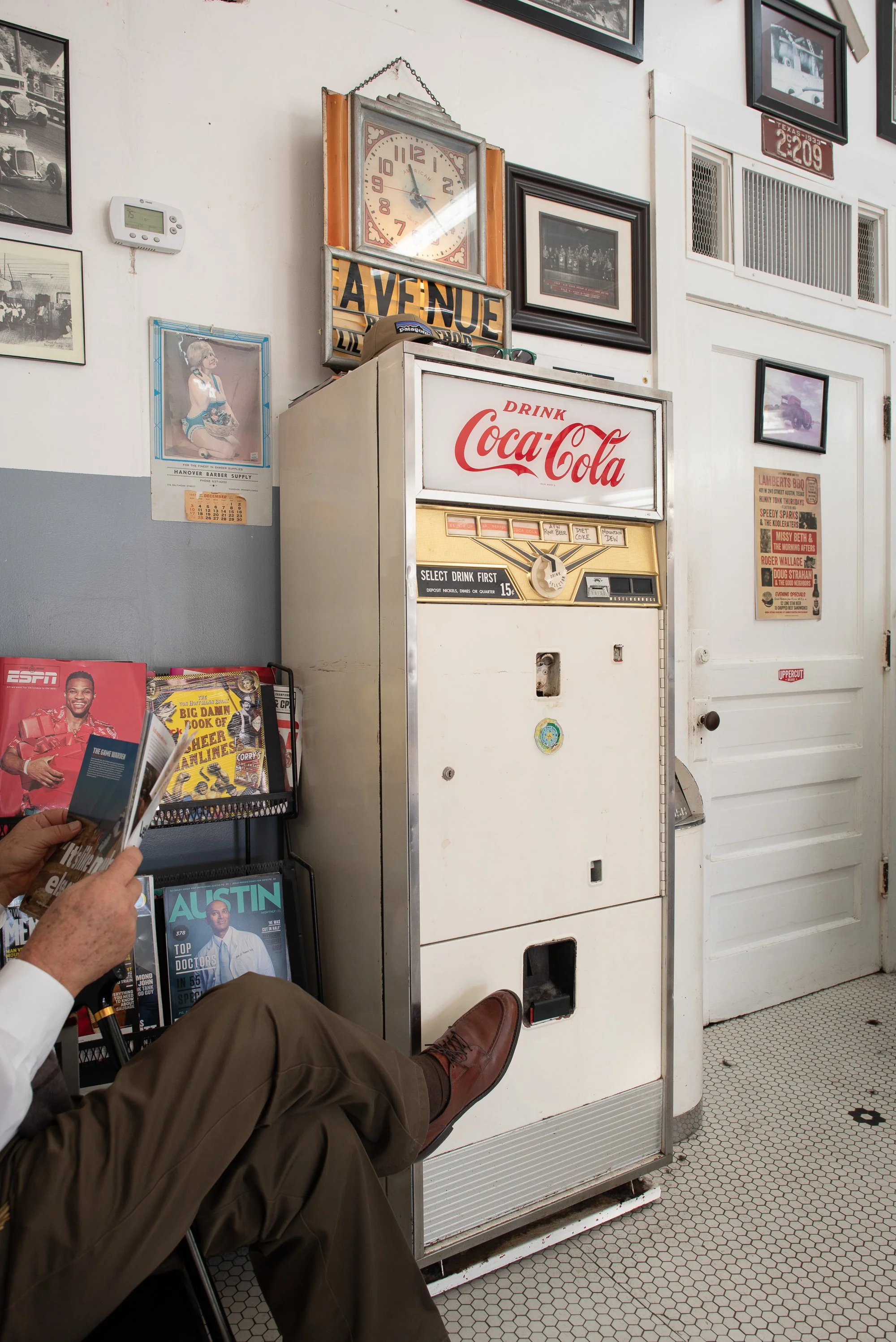 A Coca-Cola machine stands against the wall beside framed photos and posters inside Avenue Barbershop in Austin, Texas
