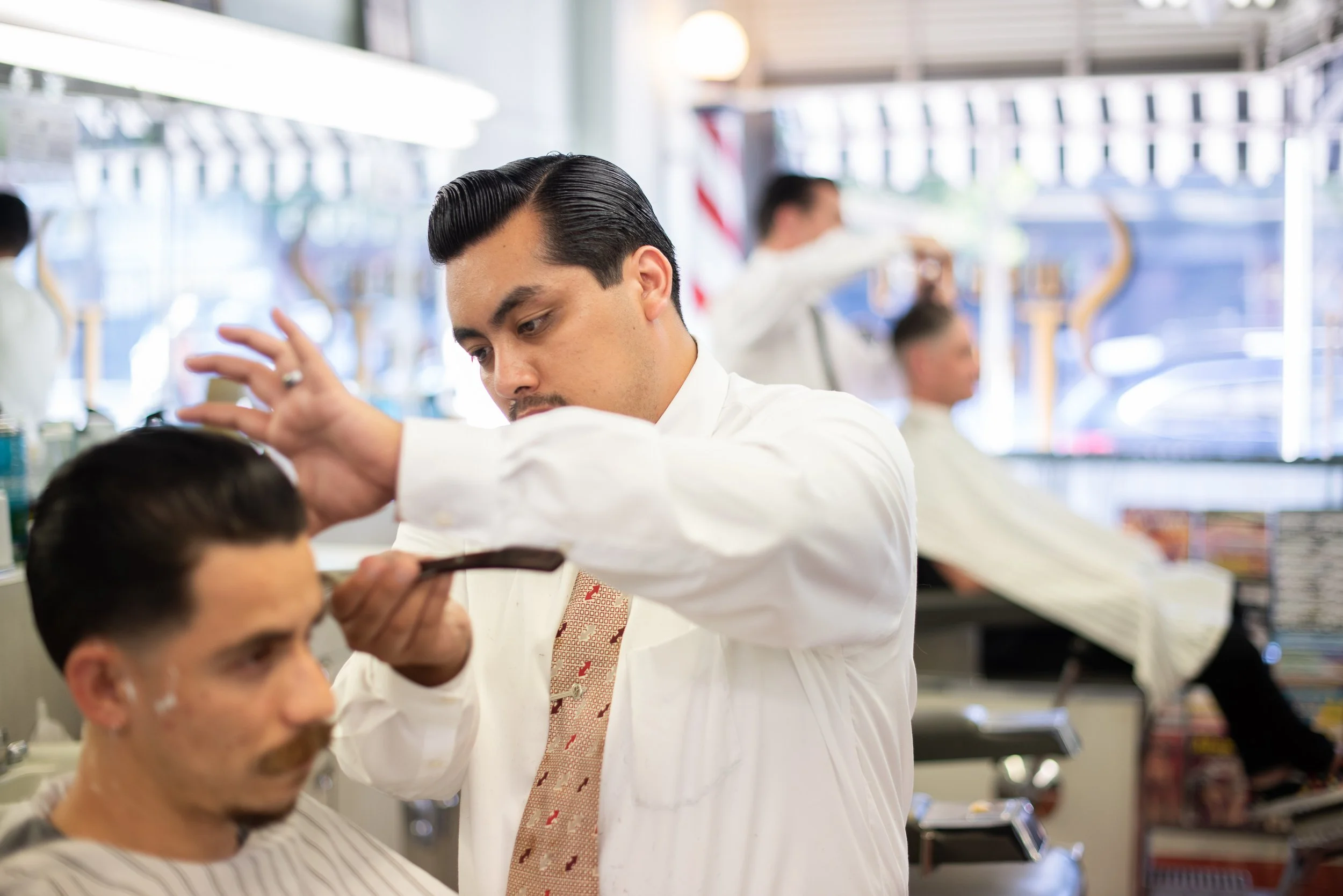 Barber in white shirt and tie working a straight razor along a client's hairline at Sweeney Todd's Barbershop, second barber and client visible in background