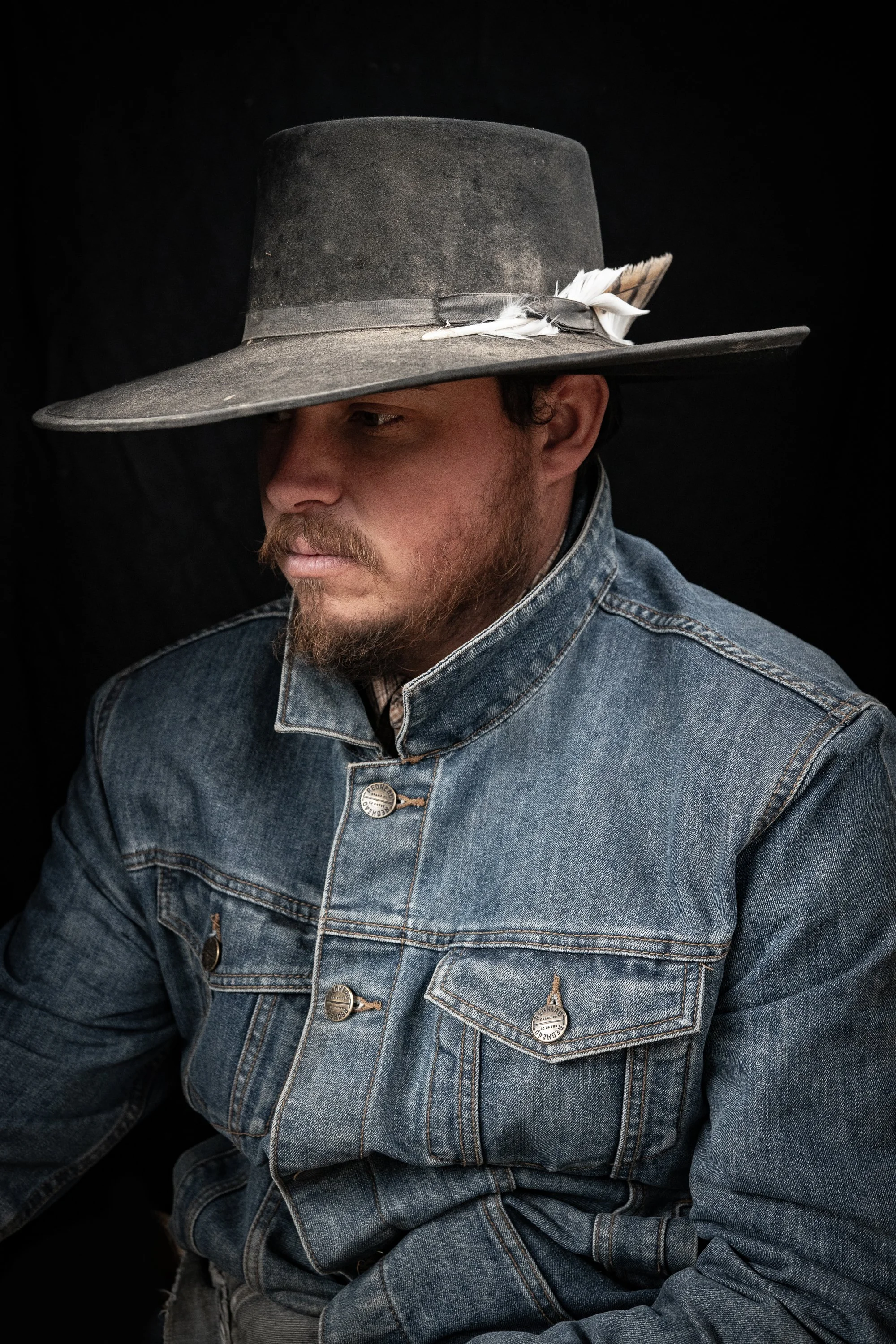 Portrait of a buckaroo wearing a weathered cowboy hat decorated with white feathers and a denim jacket against a dark background.