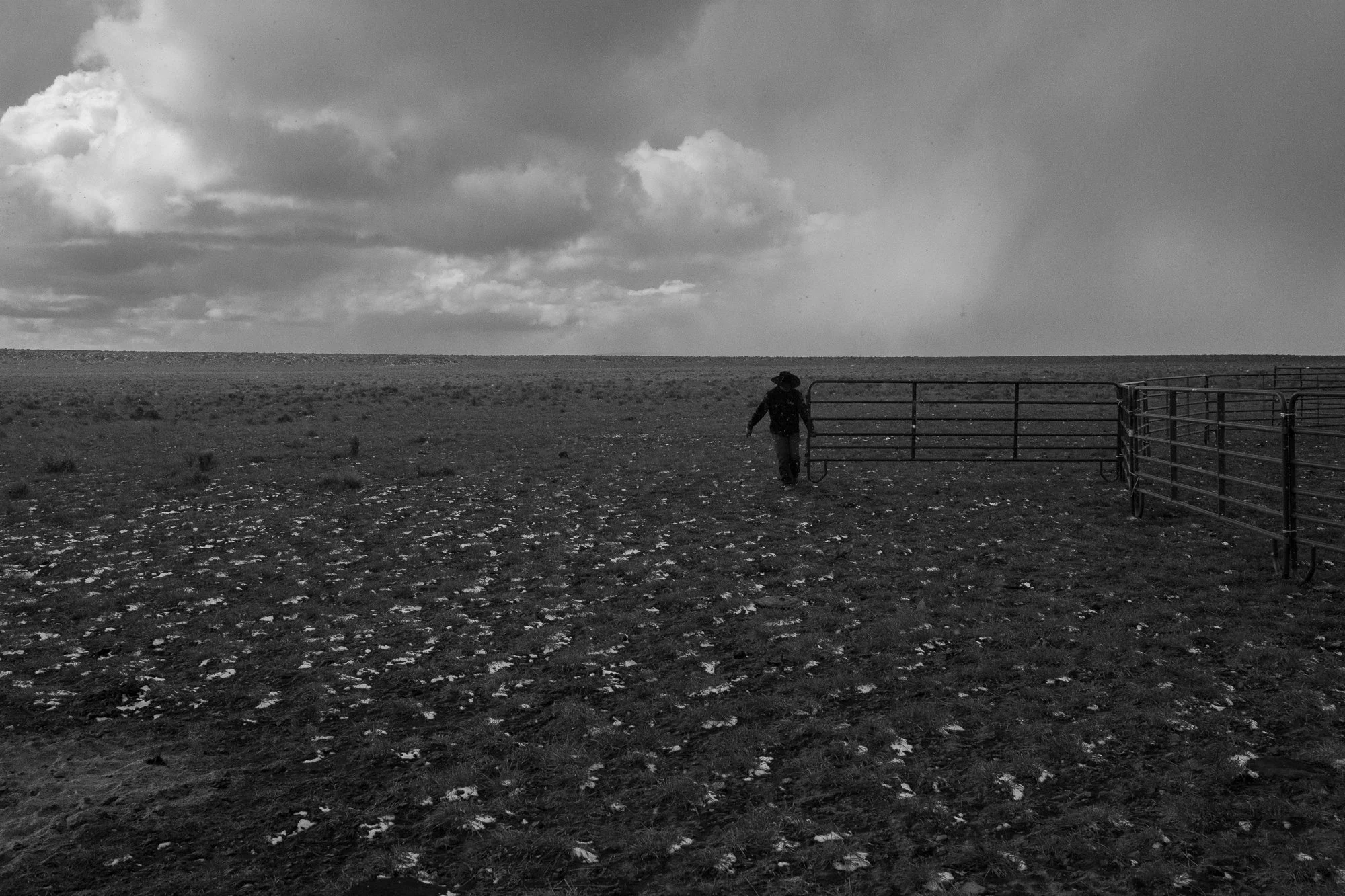 A cowboy walks near a metal gate beneath storm clouds on open ranch country at the ZX Ranch