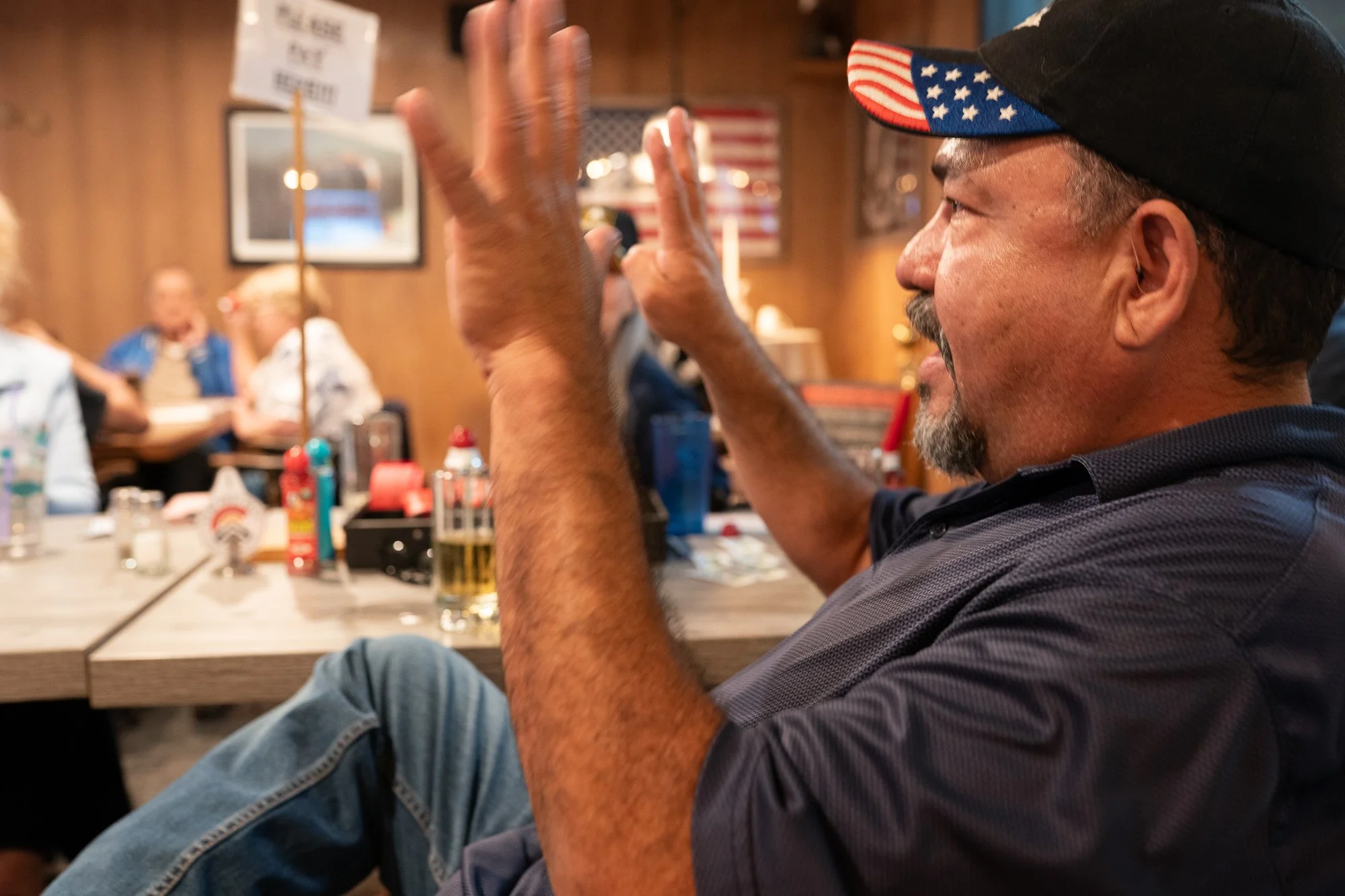 Veteran wearing an American flag brim hat speaking with his hands inside a VFW hall