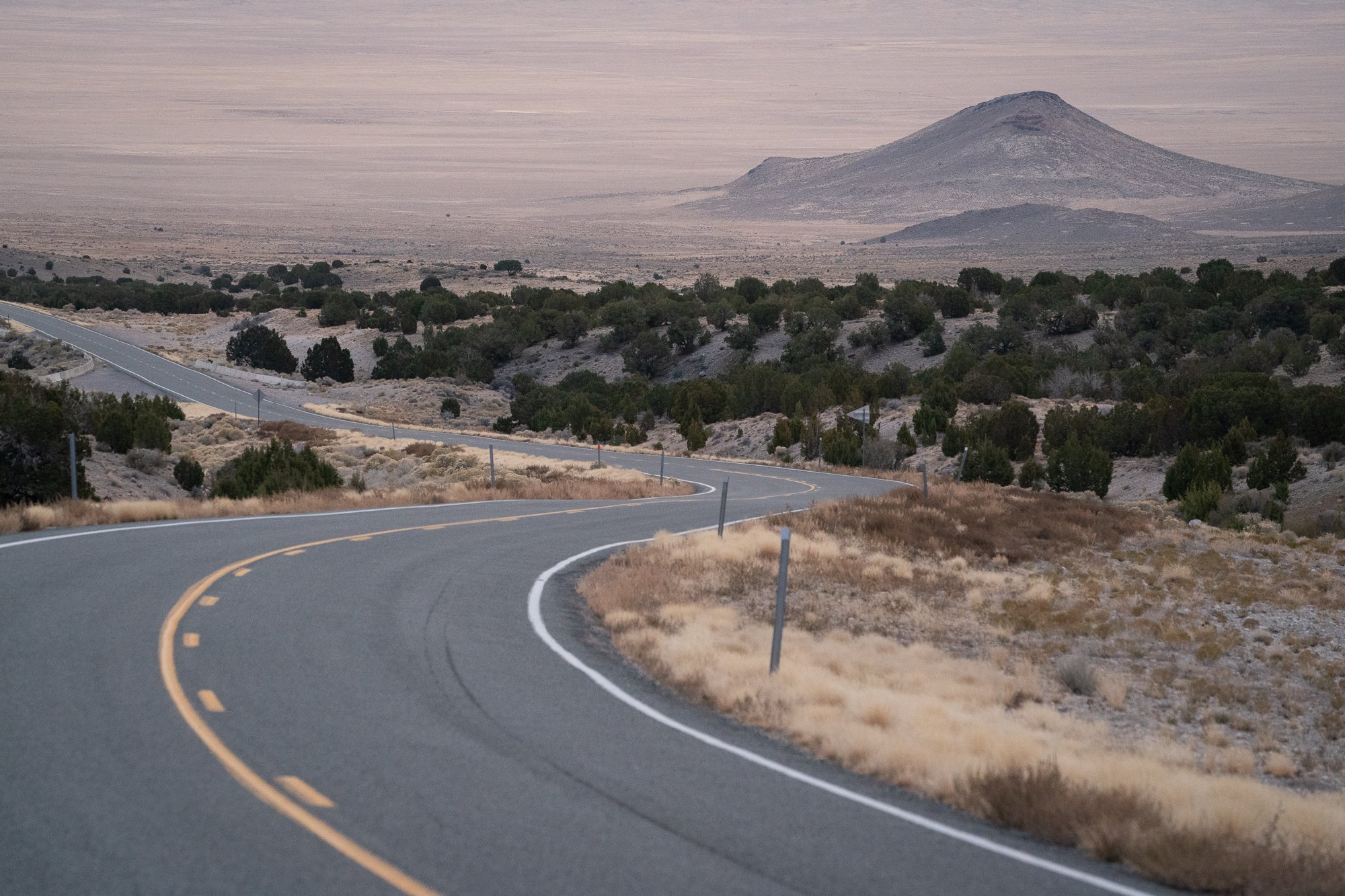 Paved two-lane road curving through low, dry vegetation toward a mountain on the horizon.