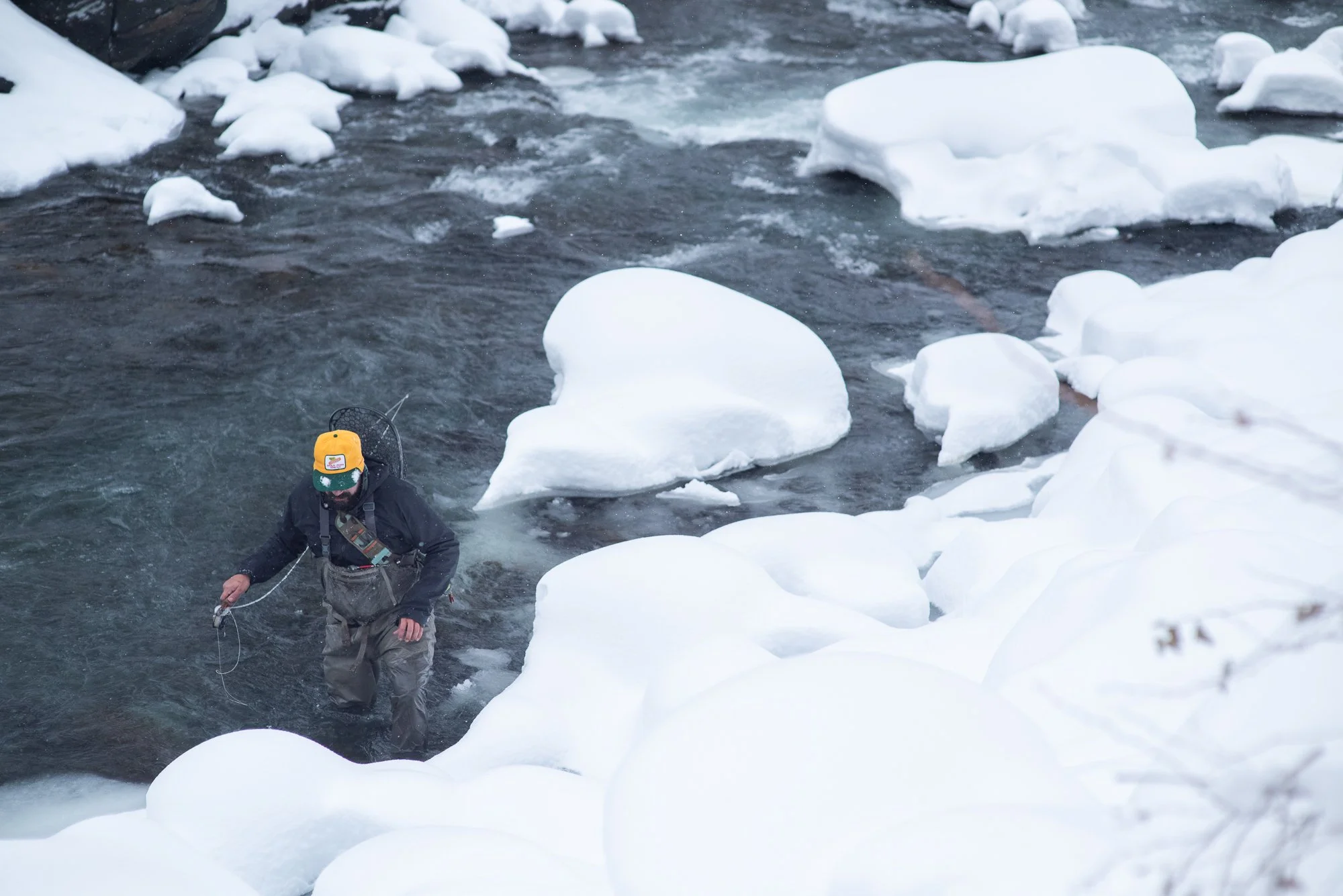 A fisherman wades through Clear Creek in Colorado during winter surrounded by snow-covered rocks and flowing water.