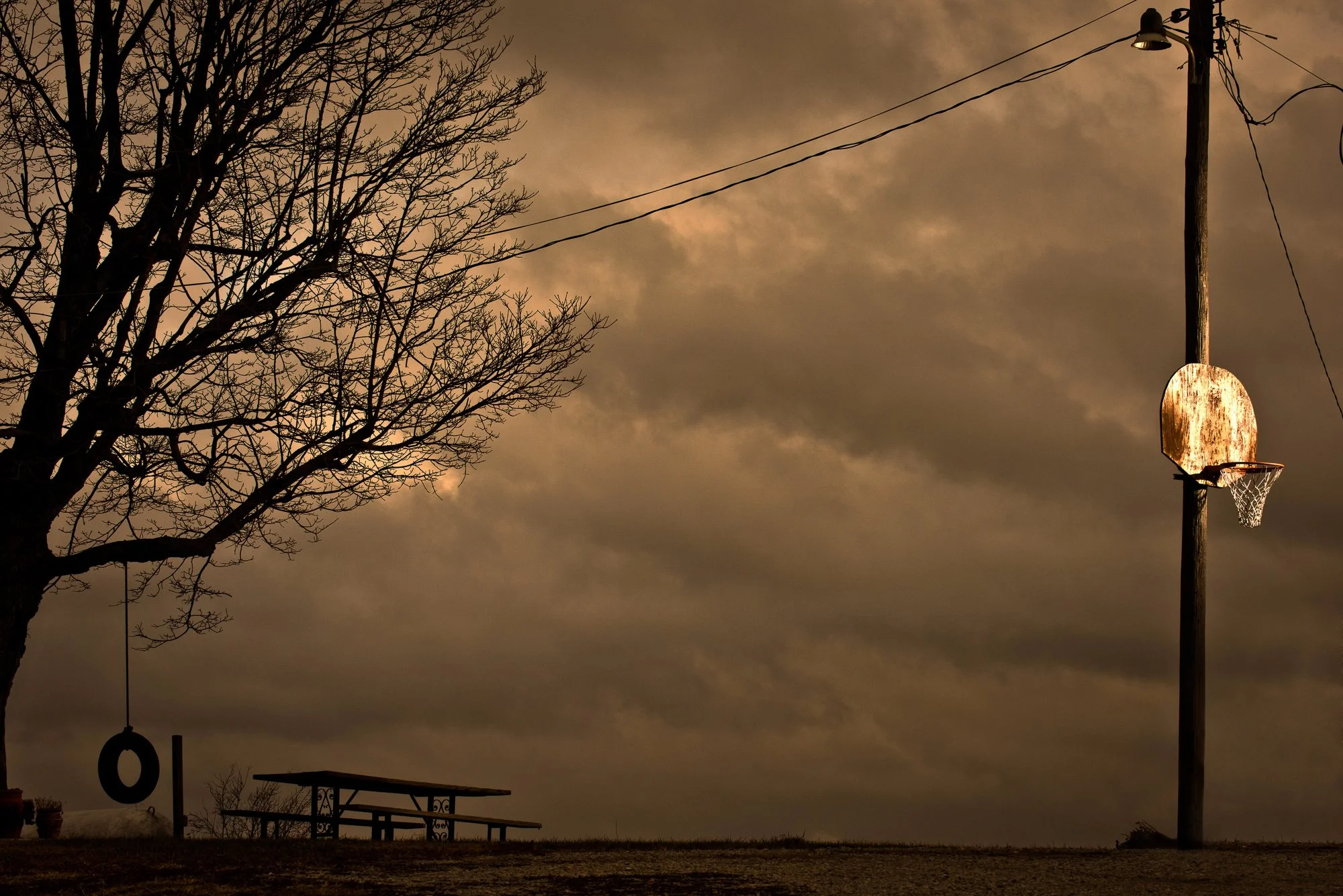 Basketball hoop beside a picnic table beneath a cloudy evening sky, with a large tree and utility pole nearby.