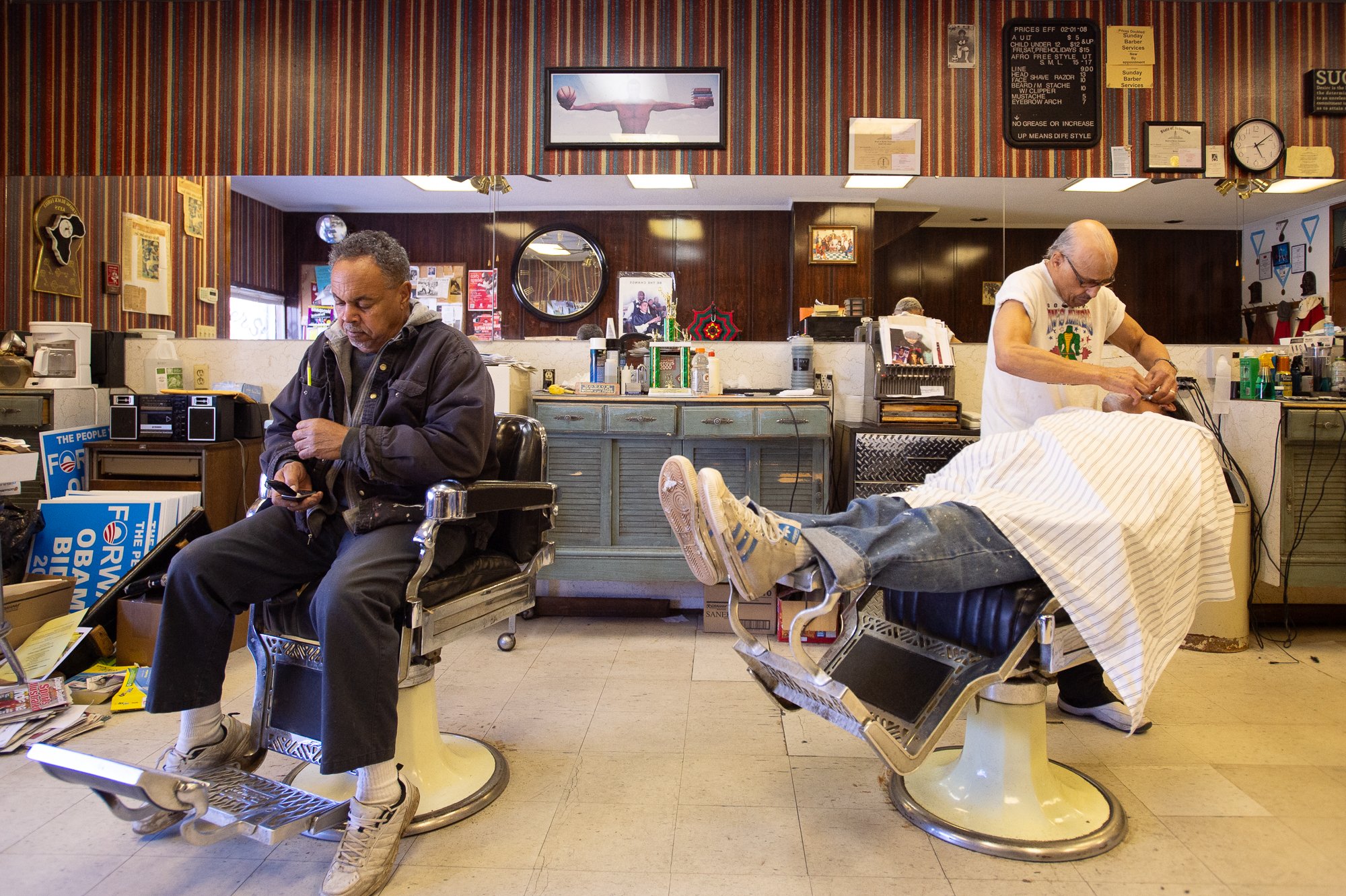 Barber cutting a client’s hair while another man sits nearby in a traditional Black barbershop