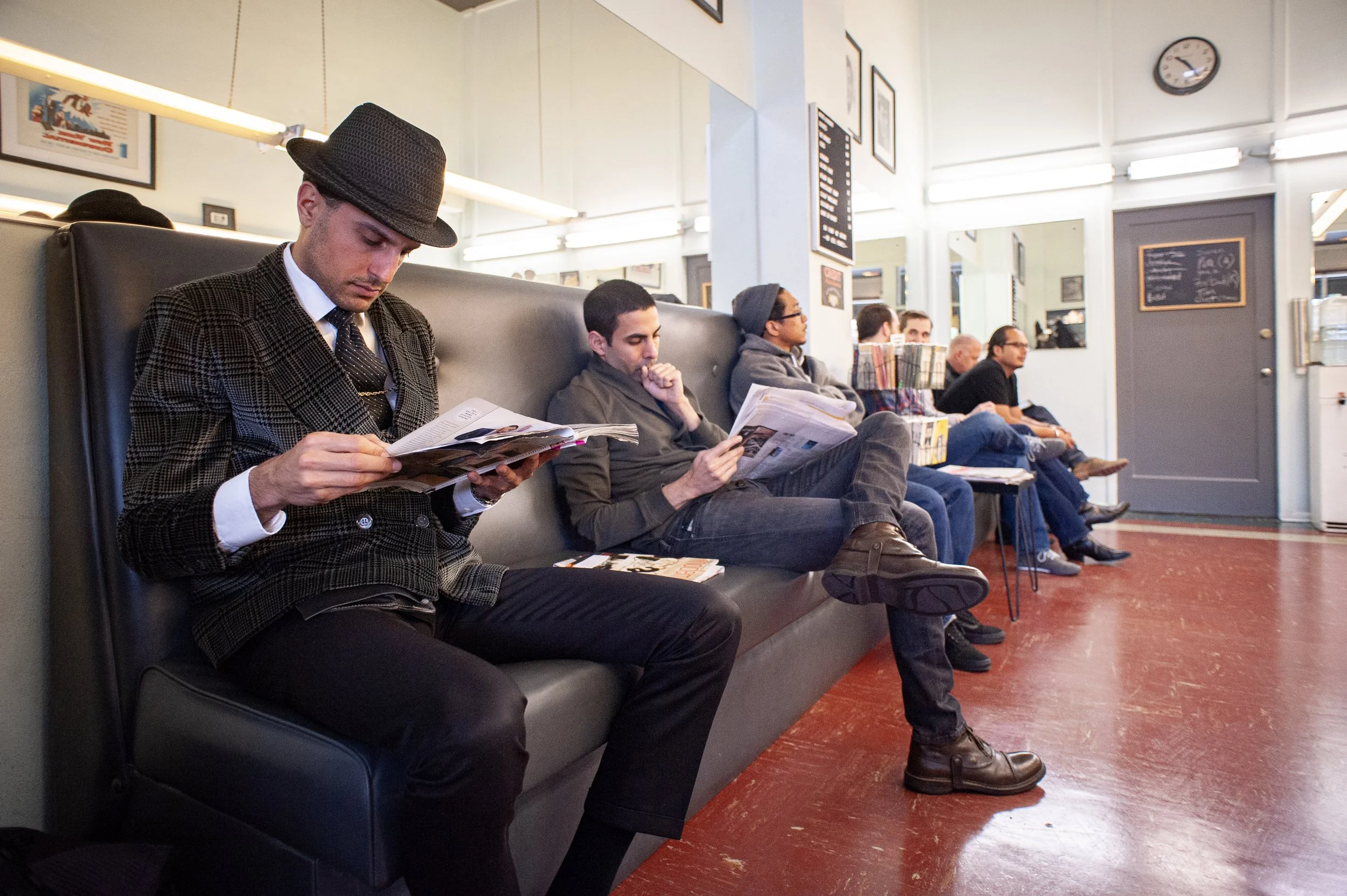 Row of men seated along a bench in Sweeney Todd's waiting area reading magazines and newspapers, with red linoleum floor, wall clock, and framed photographs behind them