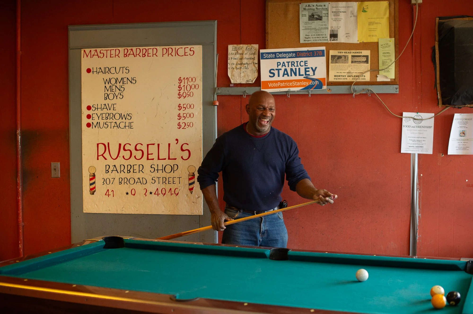 Man smiling and holding a pool cue inside a barbershop with signage and price board behind him