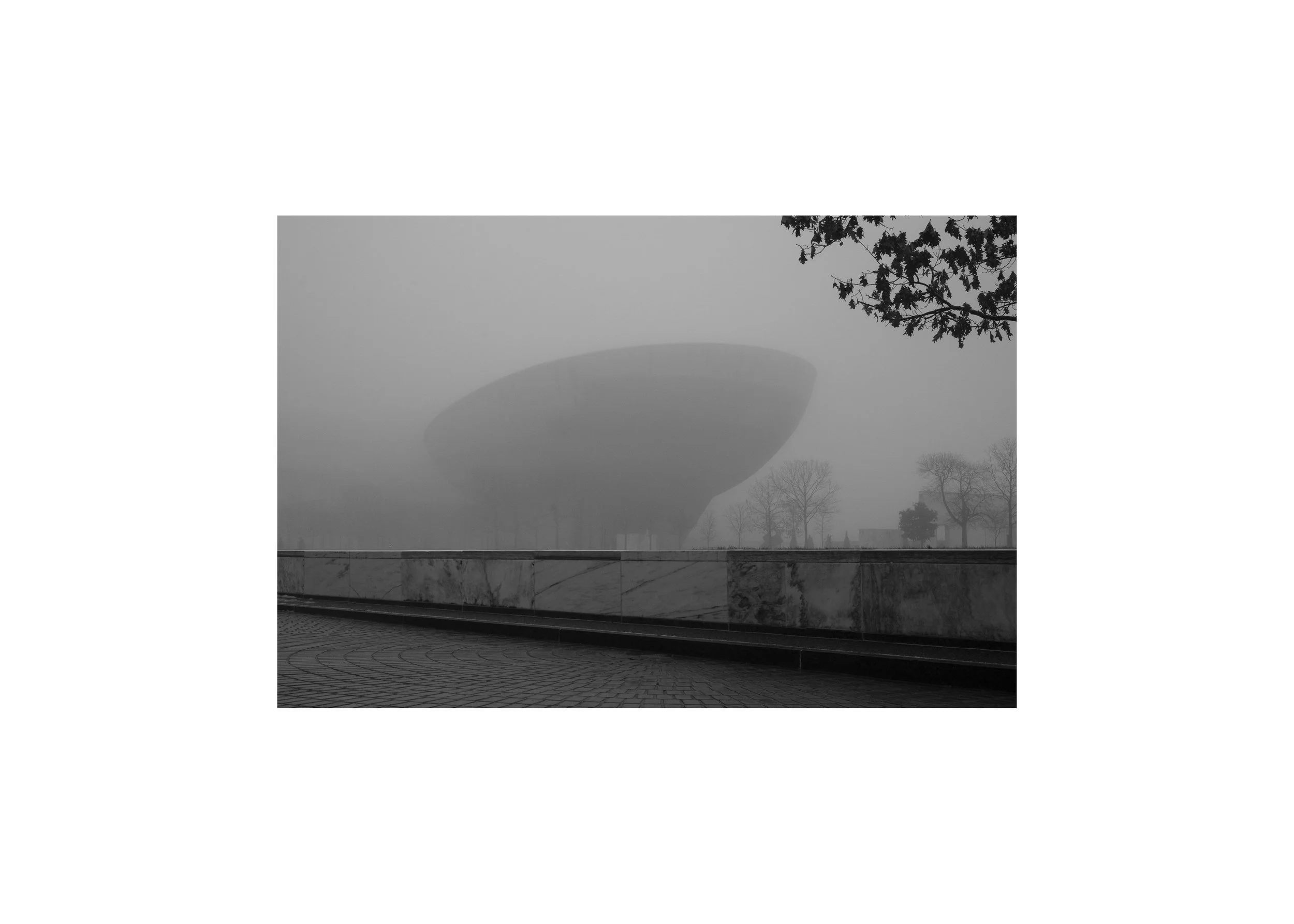 Black and white fine art print of The Egg in Albany emerging from dense fog, with stone plaza and curved wall in foreground